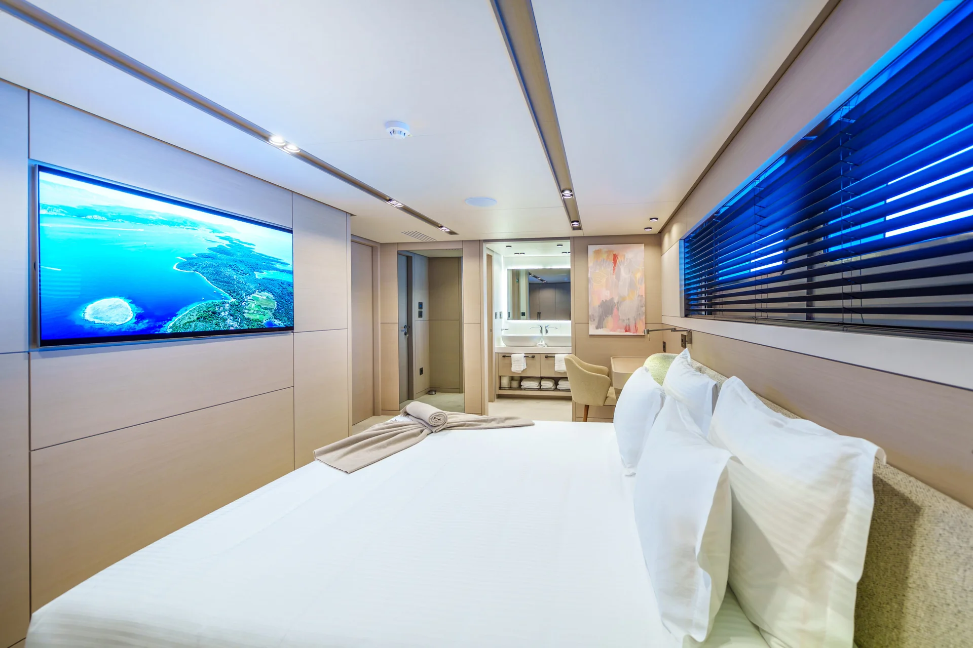 ORIY Modern yacht bedroom with a neatly made bed, white linens, large wall-mounted TV showing an aerial island view, folded towels, large window with blinds, and an ensuite bathroom visible through an open door.