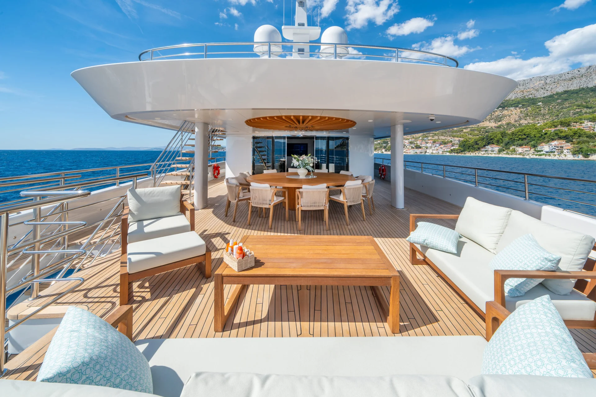 ORIY Luxury yacht deck with cushioned seating, a wooden dining table, and ocean views. Bright sunlight, clear blue sky, and distant coastline are visible in the background.