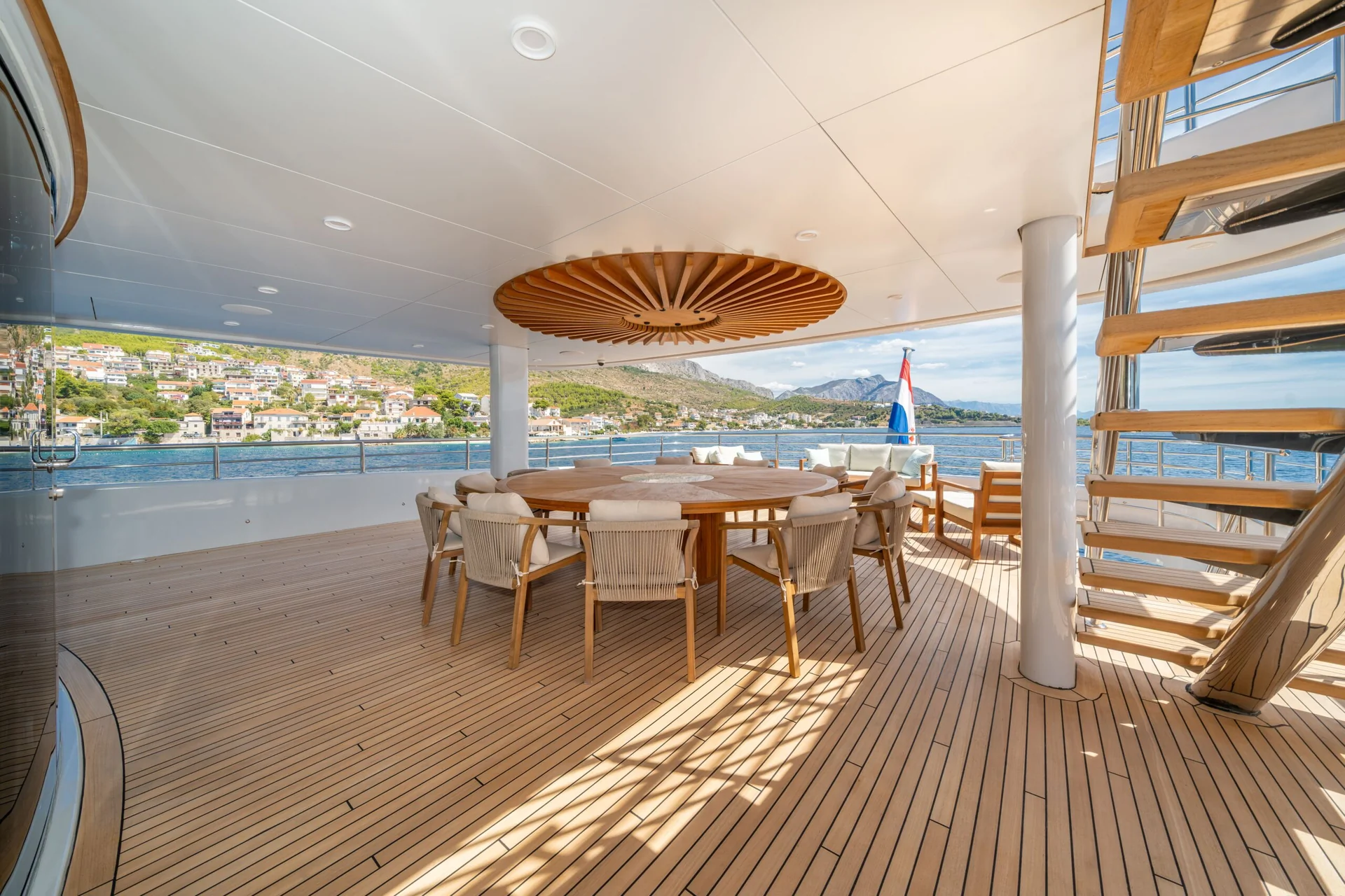 ORIY A luxurious yacht deck with a round dining table and chairs, wooden flooring, a spiral staircase, and scenic coastal views of houses and hills in the background.