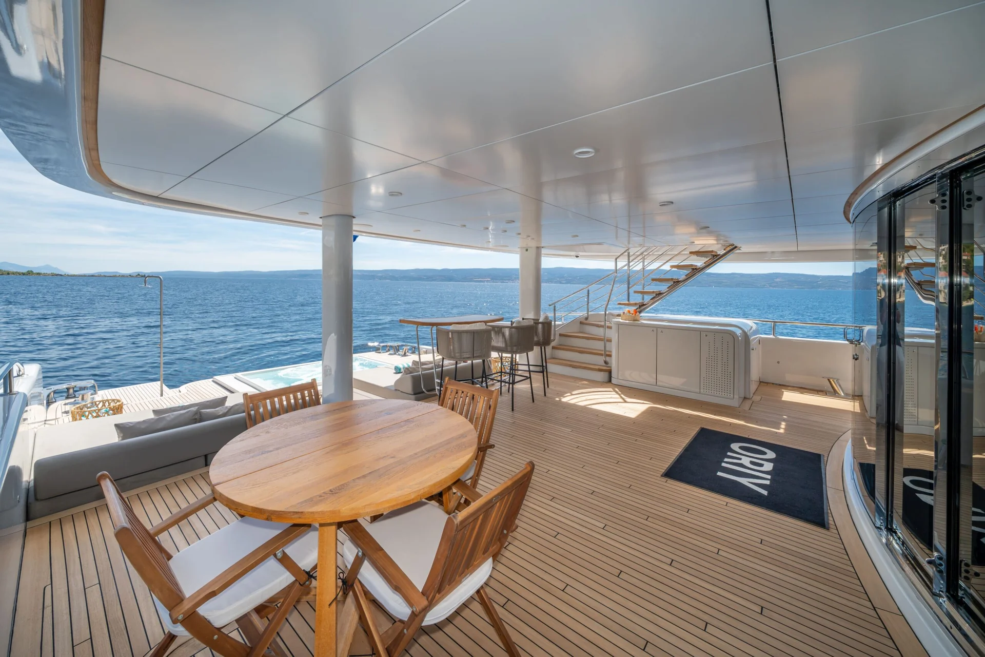 ORIY A luxury yacht deck with a round wooden table and chairs, bar seating, stairs leading up, and an ocean view. A mat by the door reads ORIY. The sky is clear and the water is calm.
