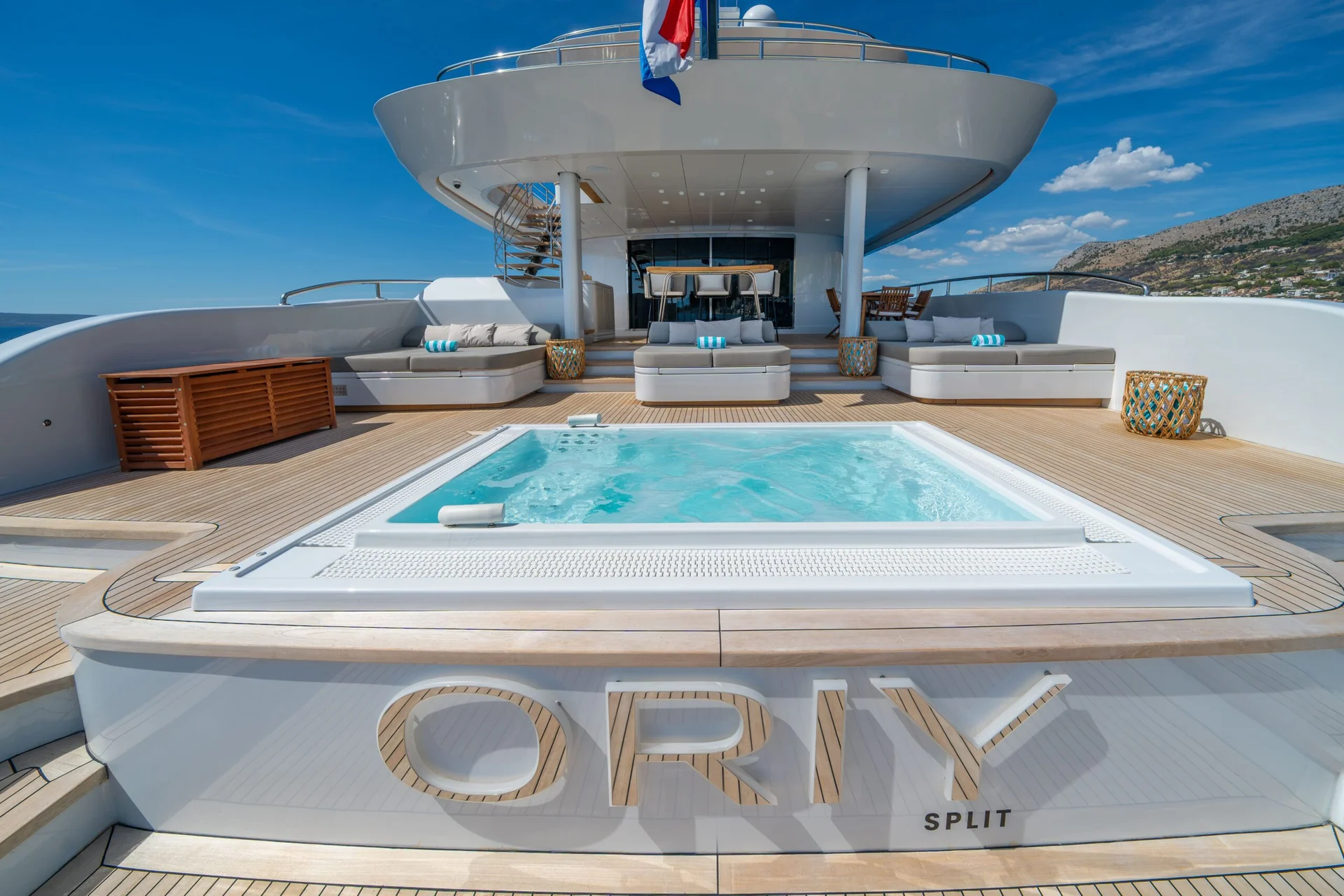 ORIY Luxury yacht deck with a jacuzzi in the center, surrounded by cushioned seating and wooden accents. The word ORIY and SPLIT appear on the yachts stern. Blue sky and coastline are visible in the background.