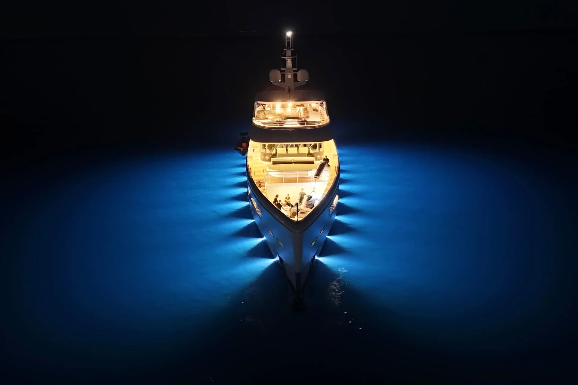 ORIY A yacht is illuminated by bright blue underwater lights at night, creating a glowing effect on the water around the boat, seen from above.