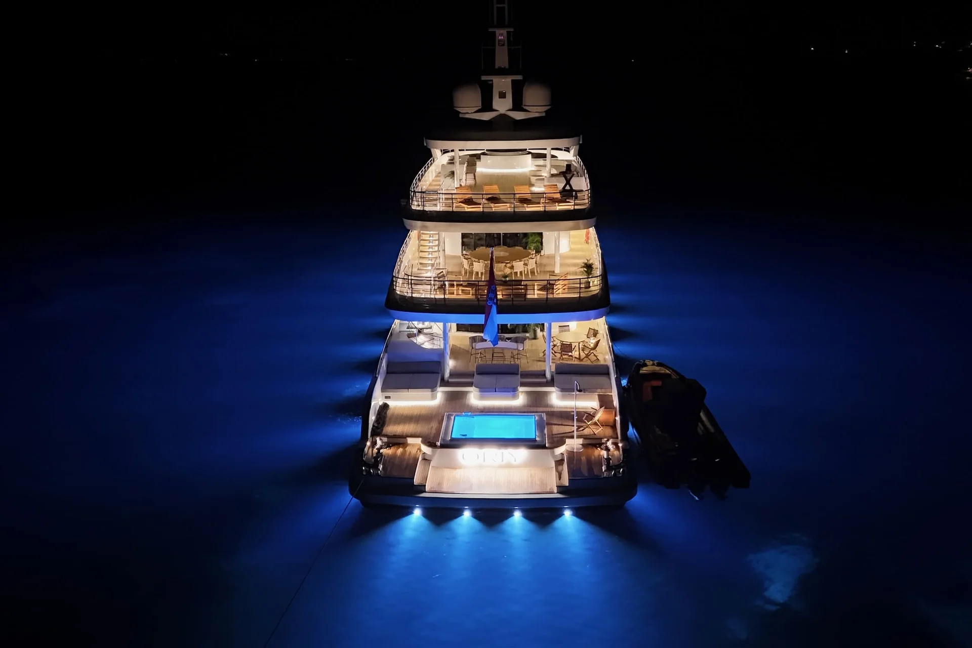 ORIY A luxury yacht at night, illuminated with blue underwater lights, showcases multiple decks, an outdoor lounge, and a pool at the stern, creating a glowing reflection on the dark water.