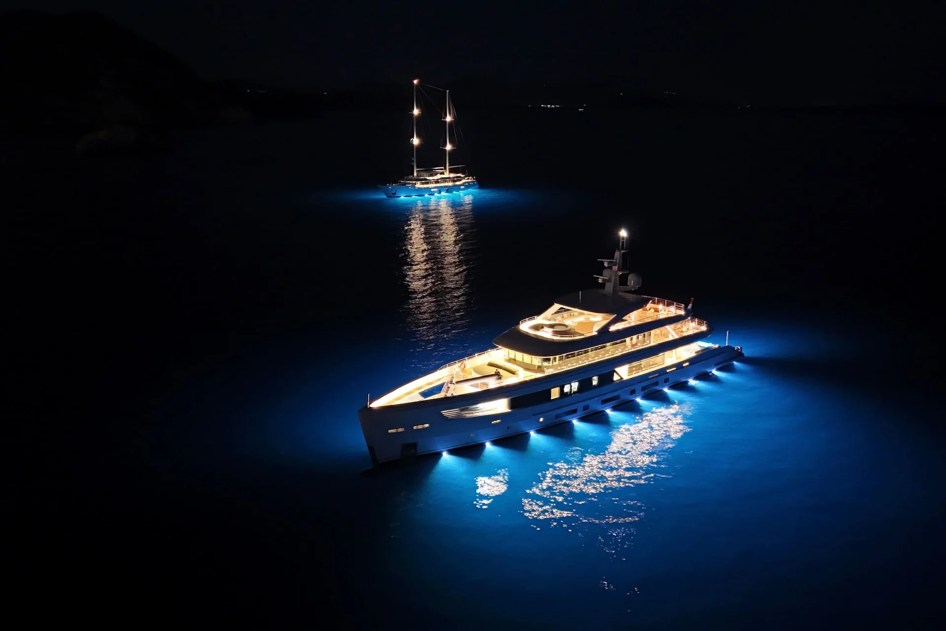 ORIY Two yachts glow with underwater blue lights on a dark sea at night. One yacht is in the foreground, brightly lit, while the second yacht is further back, also illuminated, both casting reflections on the water.