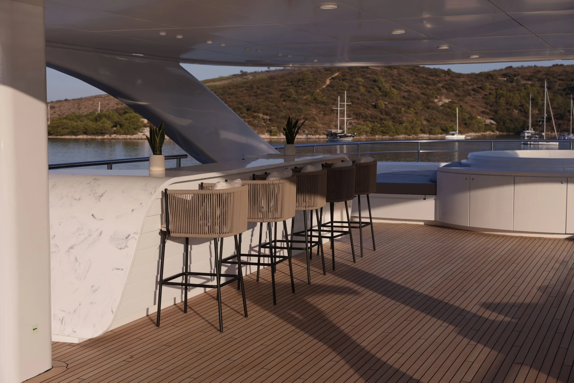 ORIY A yacht deck with a bar counter, four barstools, potted plants, and wood flooring. The deck overlooks calm water and a hilly, green coastline under a clear sky.