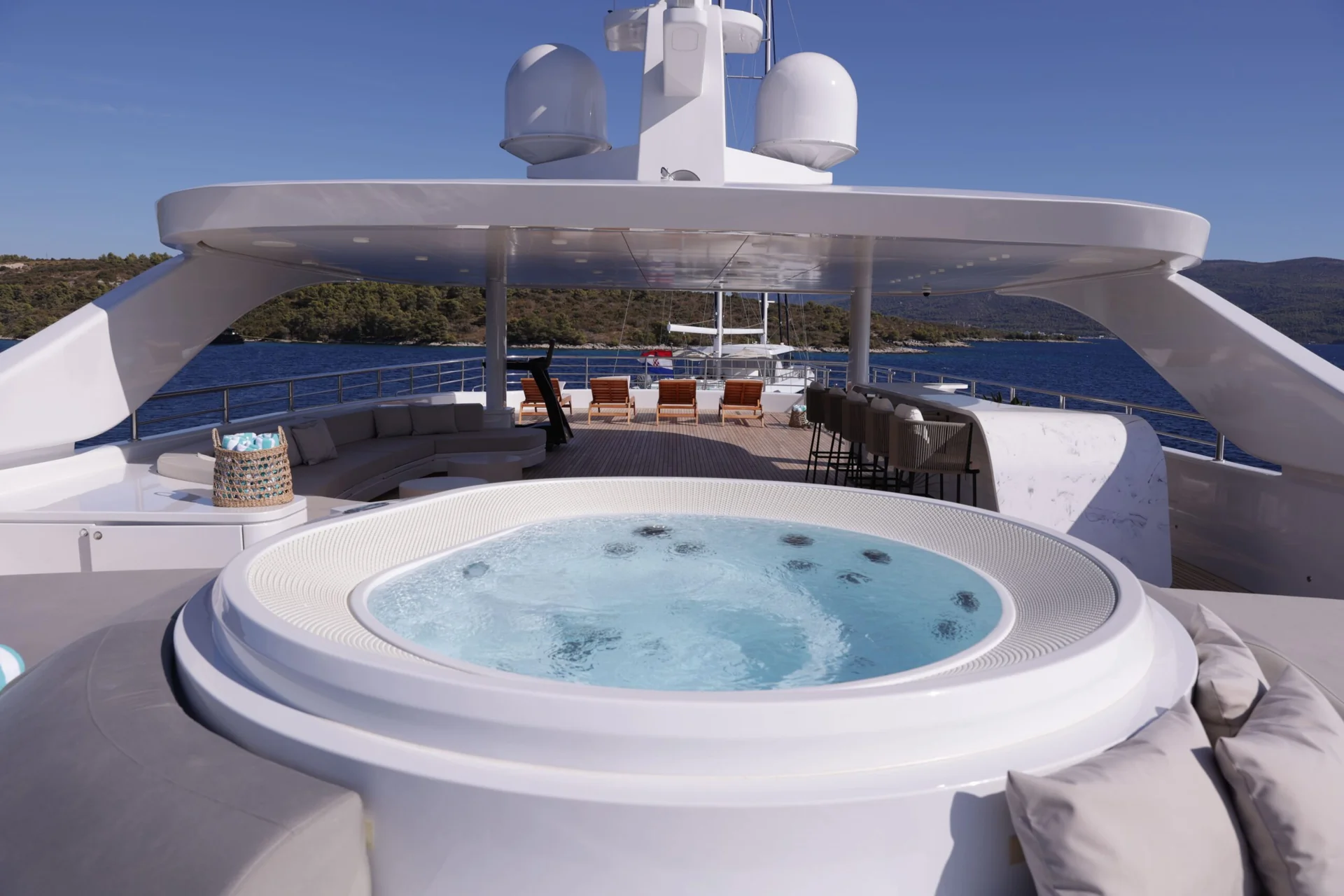 ORIY A luxurious yacht deck featuring a round hot tub in the foreground, surrounded by seating, bar stools, and open blue water under a clear sky.