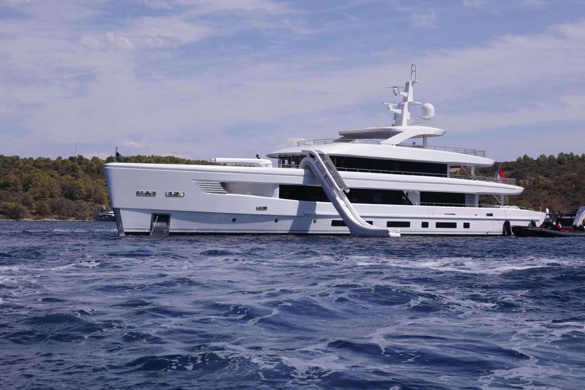 ORIY A large white yacht anchored in blue water with a long slide attached from the upper deck to the sea; a forested shoreline is visible in the background under a partly cloudy sky.