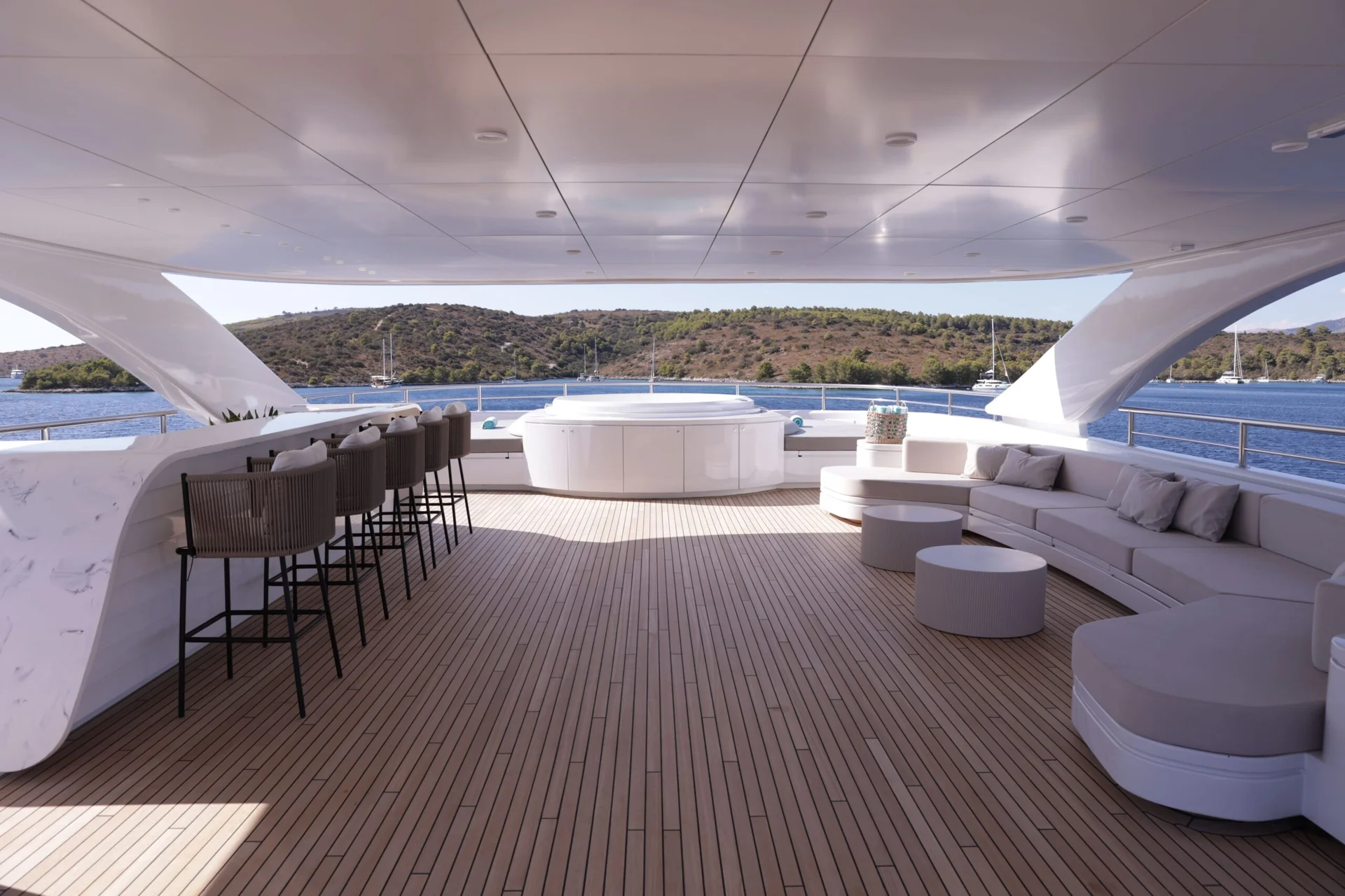 ORIY Spacious yacht deck with bar stools, lounge seating, and a hot tub, overlooking calm blue water and a hilly shoreline under a covered roof.