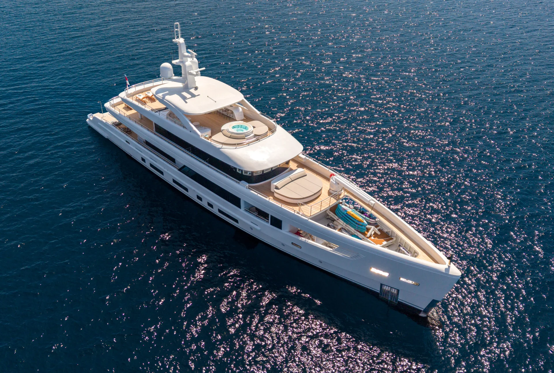 ORIY A large white luxury yacht floats on calm, sparkling blue ocean water, viewed from above. The yacht has multiple decks, outdoor seating areas, and water toys on the deck.