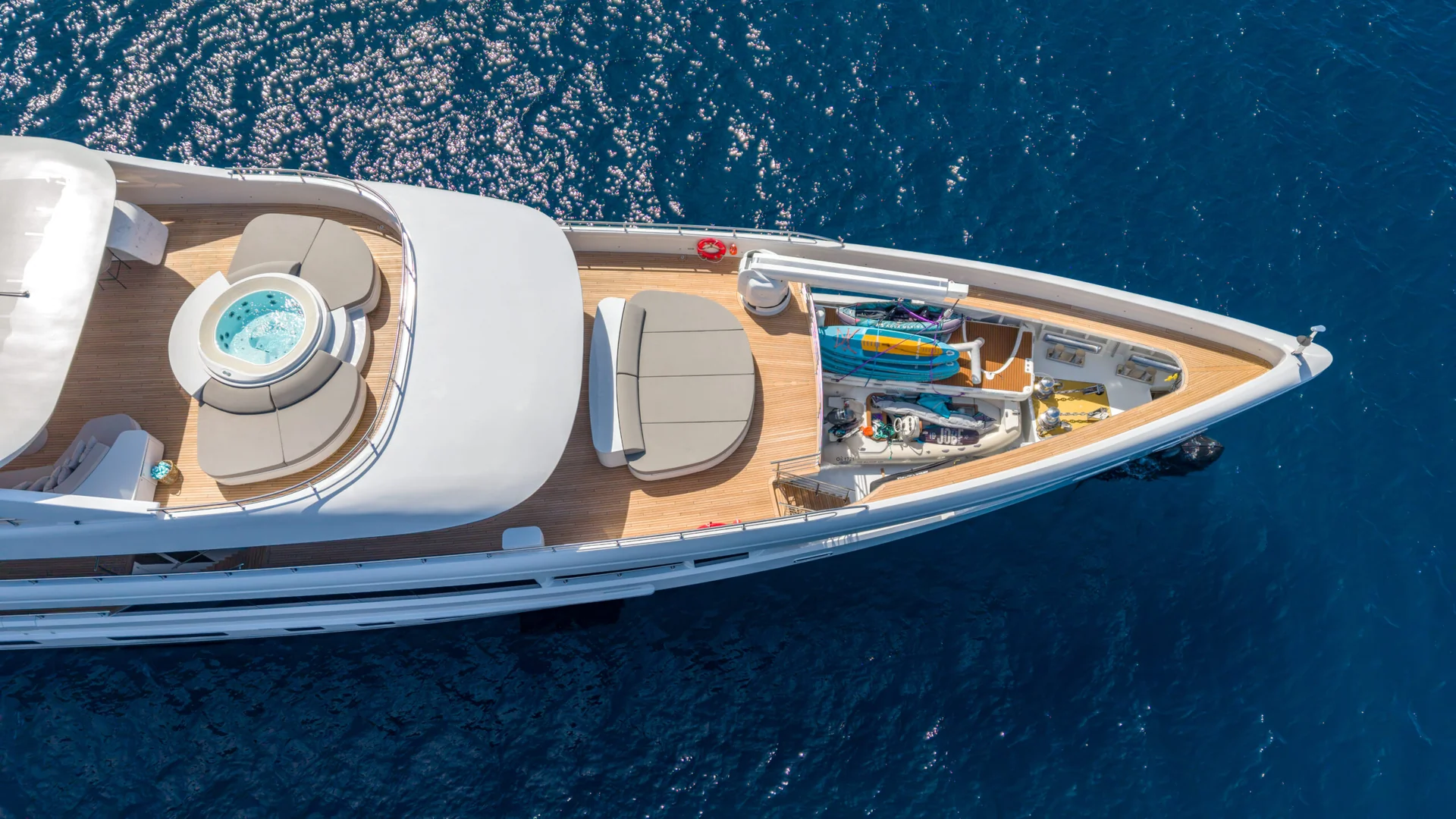 ORIY Aerial view of a luxury yachts deck, featuring lounge chairs, a round jacuzzi, and storage with water sports equipment, floating on deep blue water.