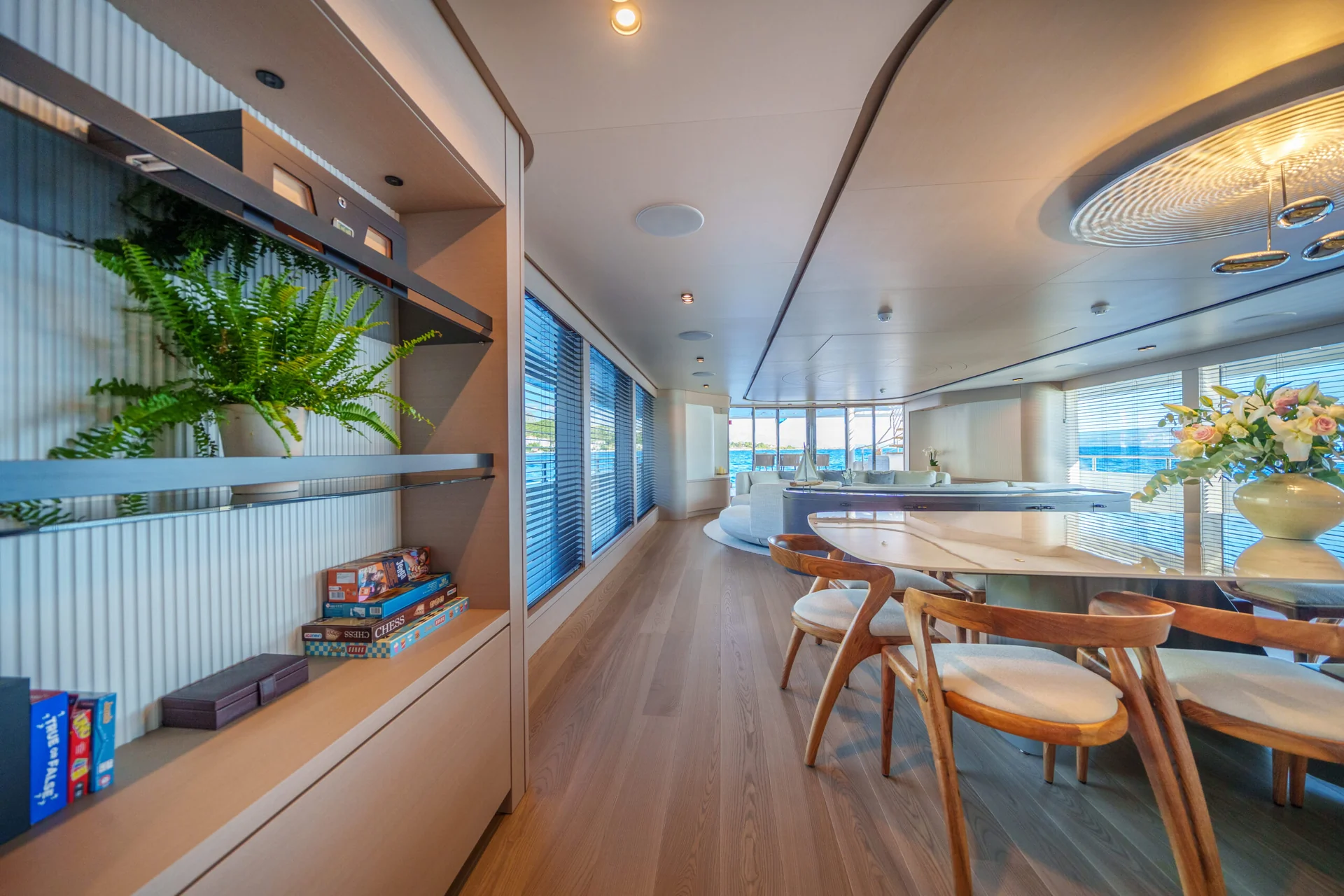 ORIY Modern interior with wooden floors, round dining table with wooden chairs, shelves holding plants and board games, large windows with blinds, and a vase of flowers on the table.