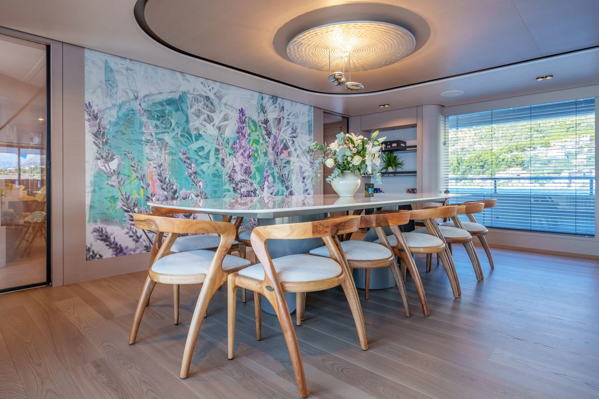 ORIY Modern dining room with a large table, ten wooden chairs with white cushions, a floral centerpiece, a colorful mural on the wall, wood floors, and large windows with blinds letting in natural light.