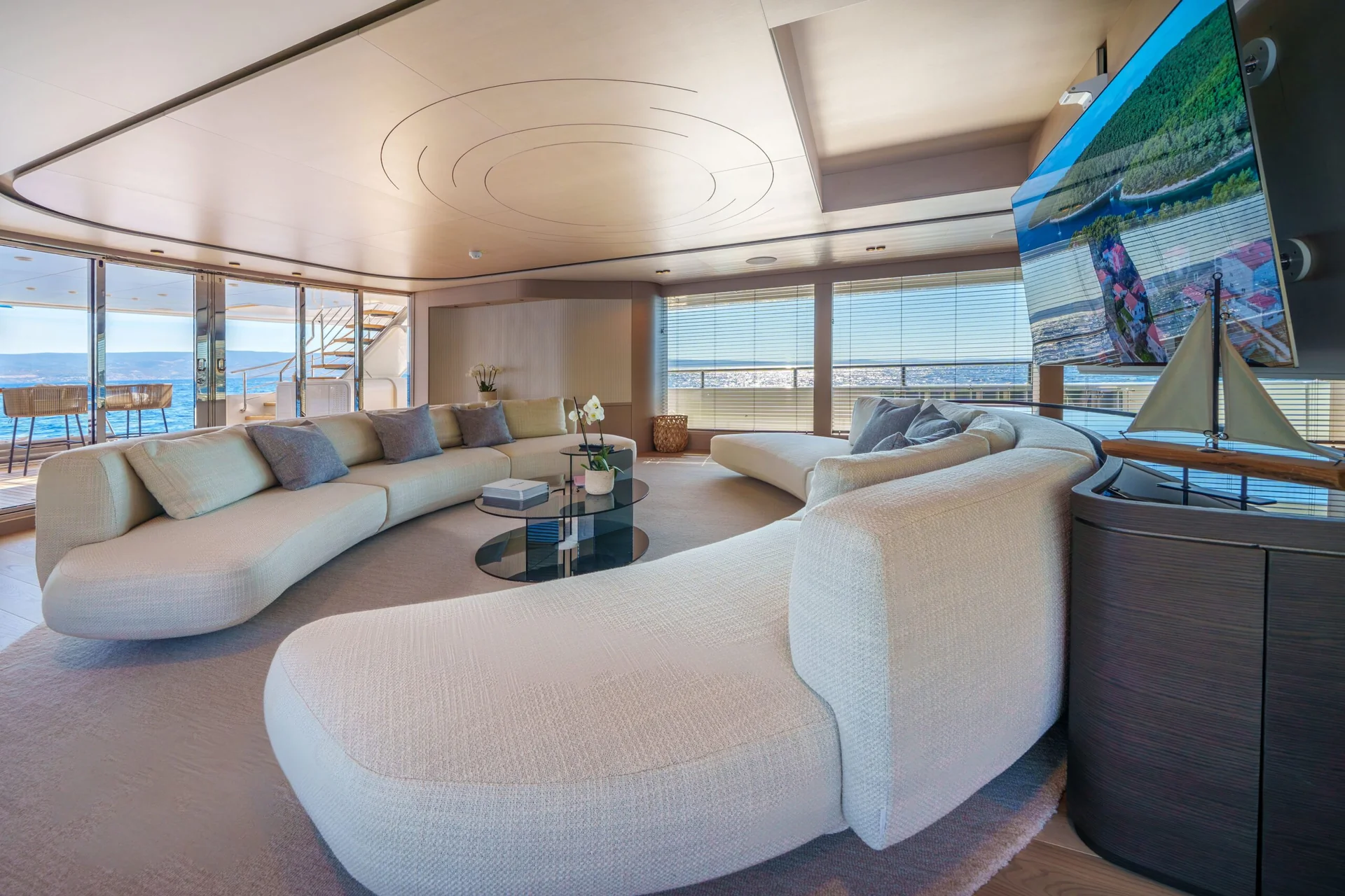 ORIY Modern living room with large curved white sofas, glass coffee tables, and a wall-mounted TV displaying nature. Floor-to-ceiling windows offer an ocean view, filling the space with natural light.