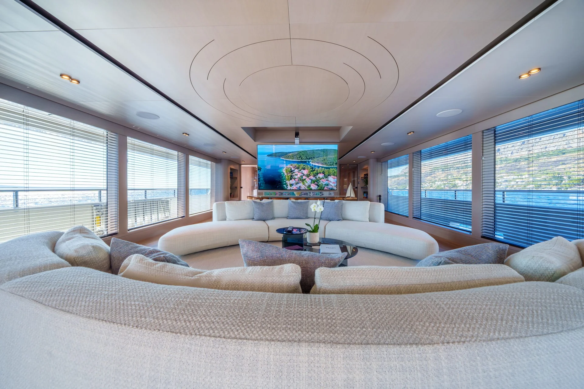 ORIY Spacious, modern yacht lounge with a curved beige sofa facing a large TV displaying a tropical landscape. Floor-to-ceiling windows offer ocean and shoreline views. A small table with flowers sits at the sofas center.