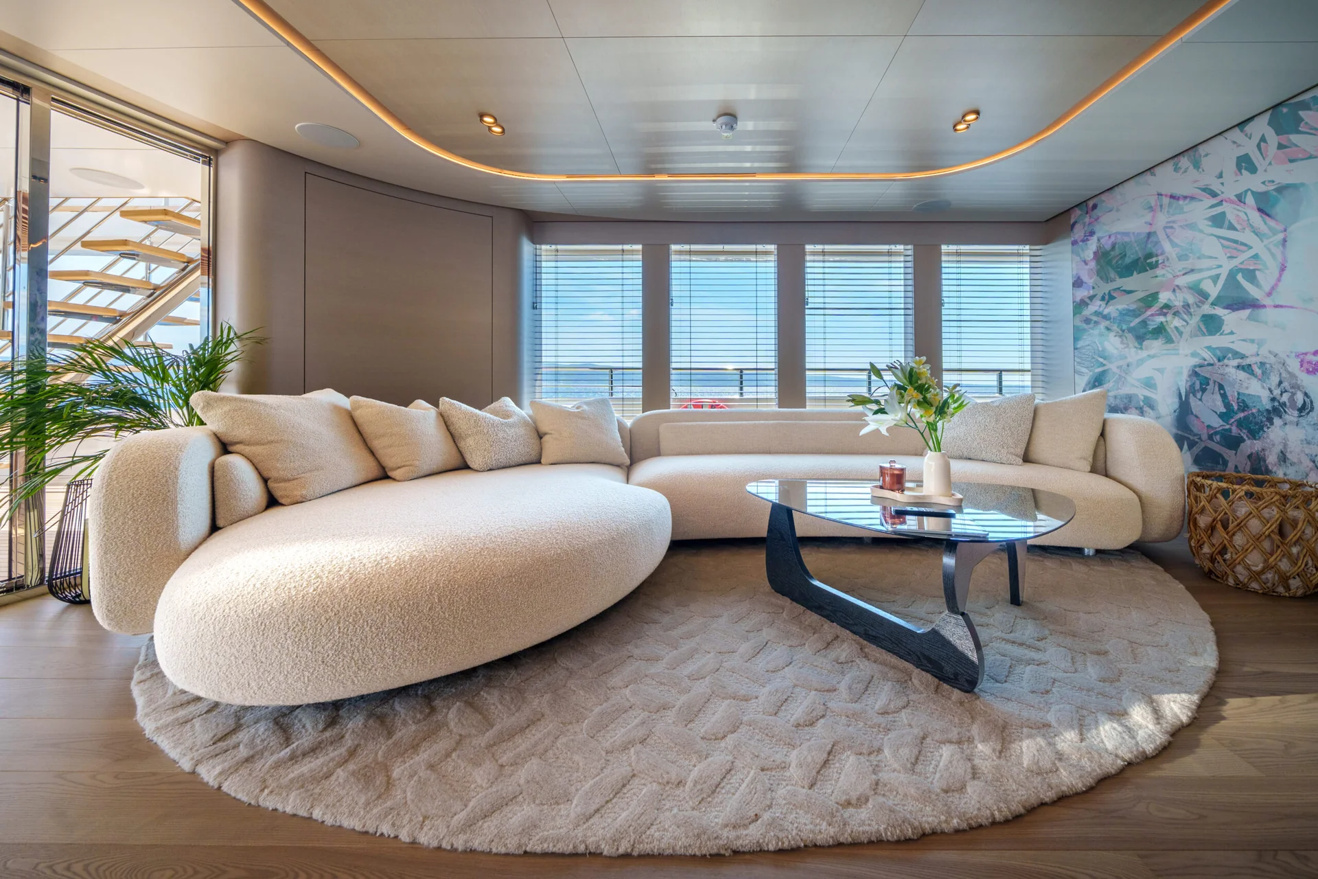 ORIY Modern living room with a large, curved cream sofa, round textured rug, coffee table with a vase of flowers, large windows with blinds, a plant, and abstract wall art. Natural light fills the space.