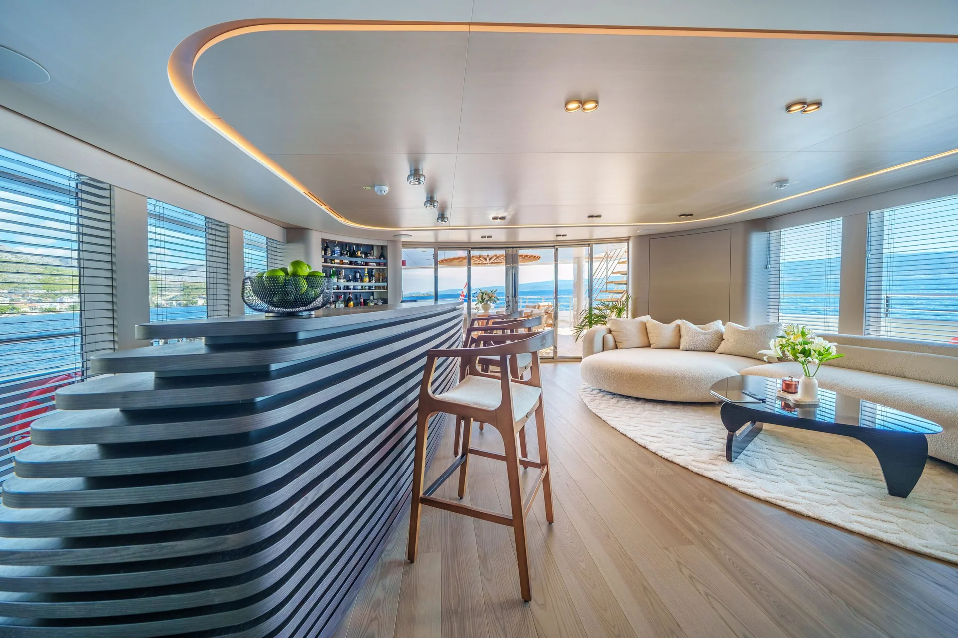 ORIY Modern yacht interior with curved bar, wooden stool, round beige sofa, large windows with blinds, and water views outside. Light wood floors and contemporary decor create a spacious, elegant atmosphere.