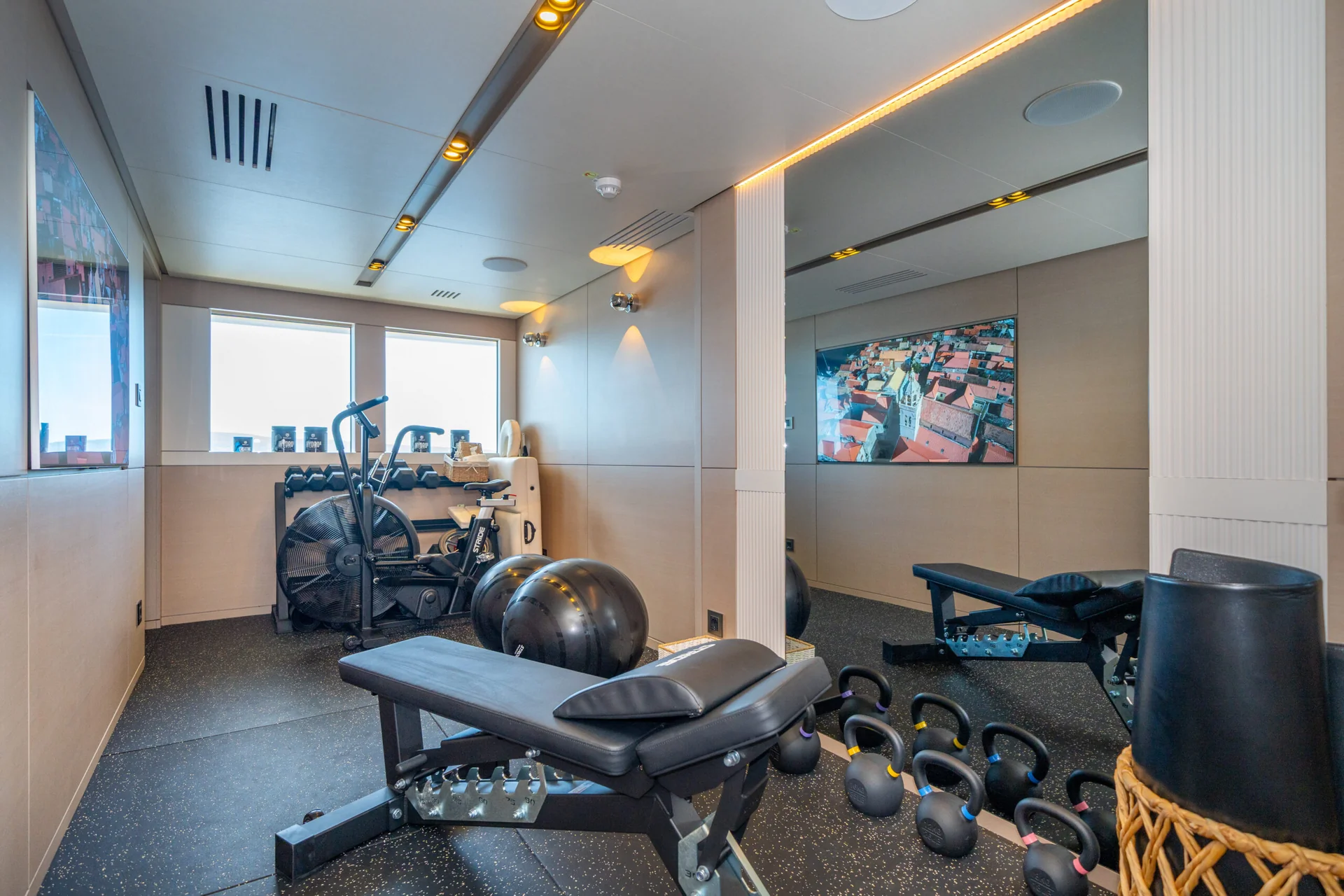 ORIY Modern gym room with a workout bench, exercise balls, kettlebells, dumbbells, and cardio machines. Large mirrors and a window provide a bright, spacious feel. A large wall photo adds decoration.