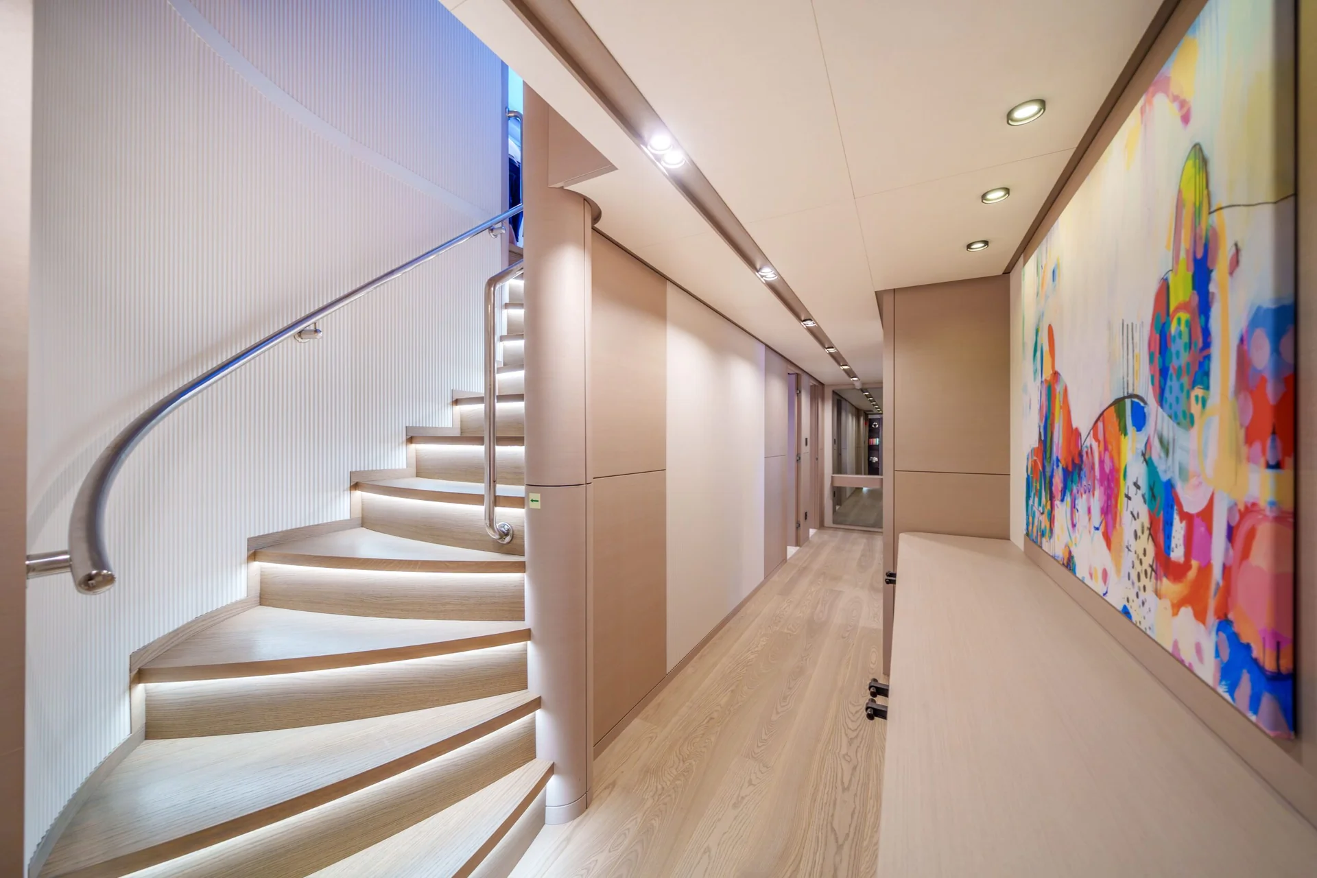 ORIY A modern hallway with light wood floors and a curved staircase with illuminated steps. Colorful abstract art hangs on the wall to the right, and recessed ceiling lights brighten the space.
