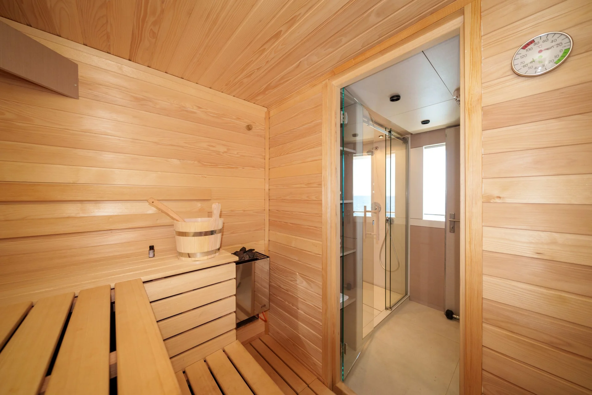 ORIY A modern wooden sauna room with benches, a water bucket, a heater, and a glass door leading to a shower area. A thermometer is mounted on the wall.