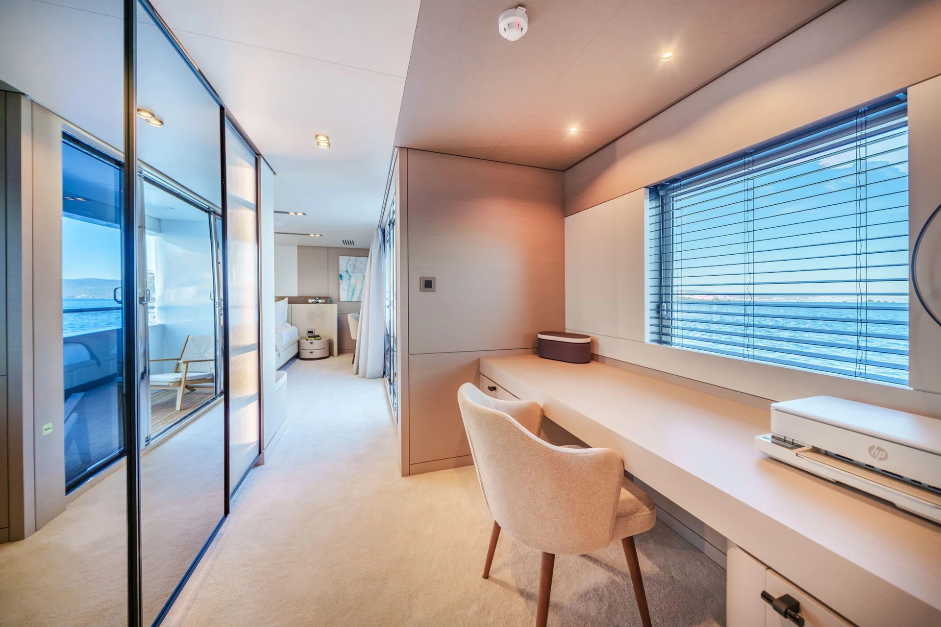 ORIY Modern yacht cabin office with a beige chair at a built-in desk, large window with blinds overlooking the ocean, glass doors, and neutral decor in soft lighting.