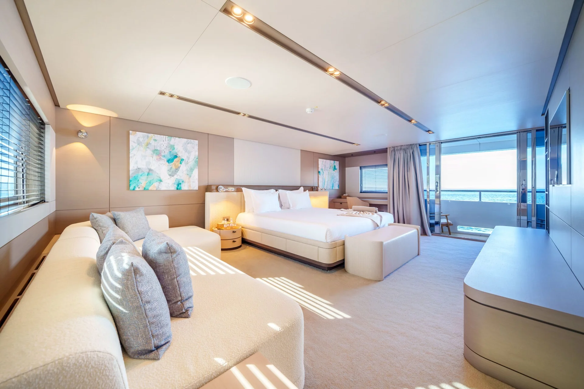 ORIY Modern, spacious bedroom with large bed, sofa, and wall art, featuring neutral tones. Sunlight streams in through windows, and glass doors open to a balcony with a view of the ocean.