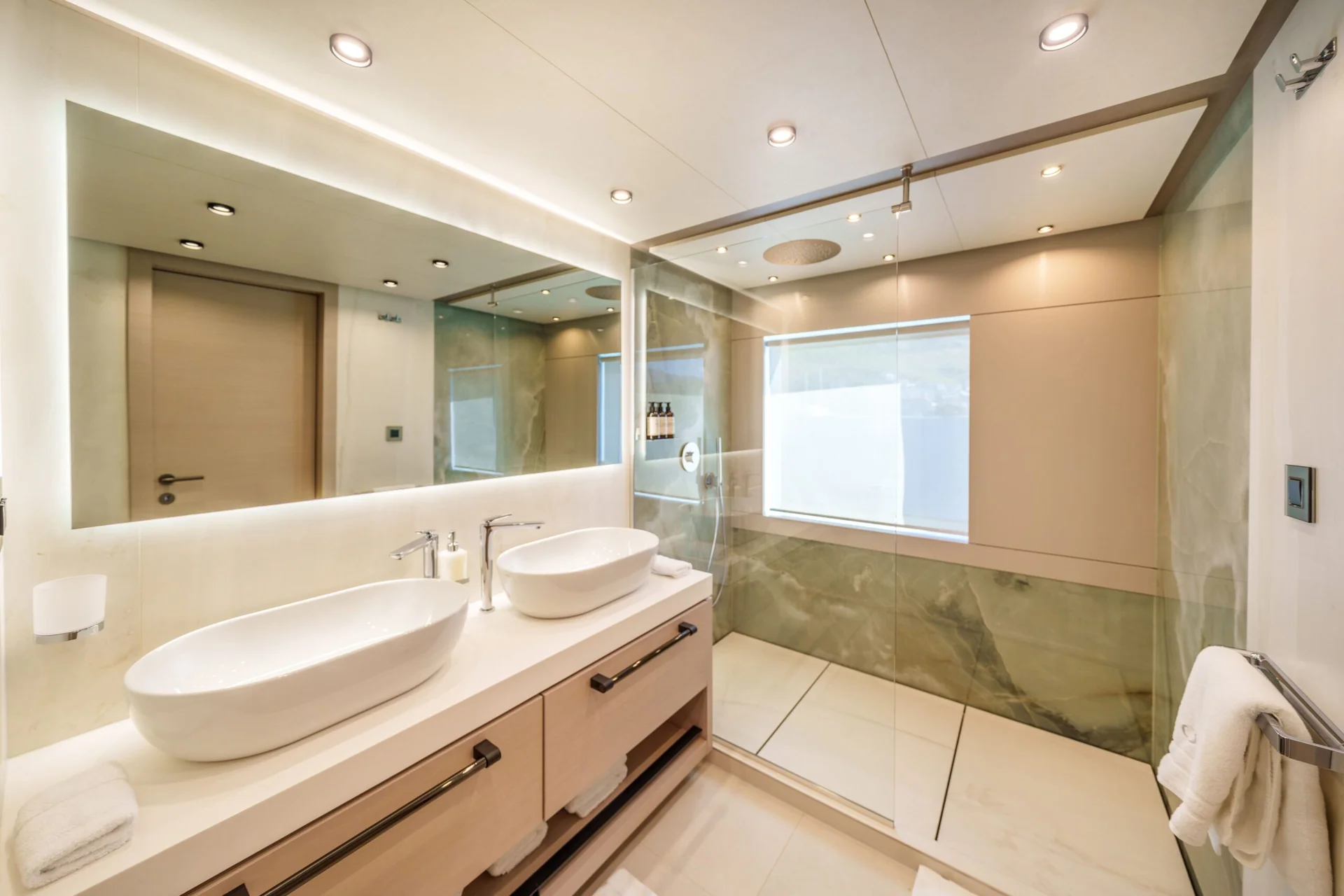ORIY Modern bathroom with a double sink vanity, large mirror, walk-in glass shower, light wood cabinetry, and recessed lighting. Towels are neatly hung, and the space is clean and bright with neutral tones.