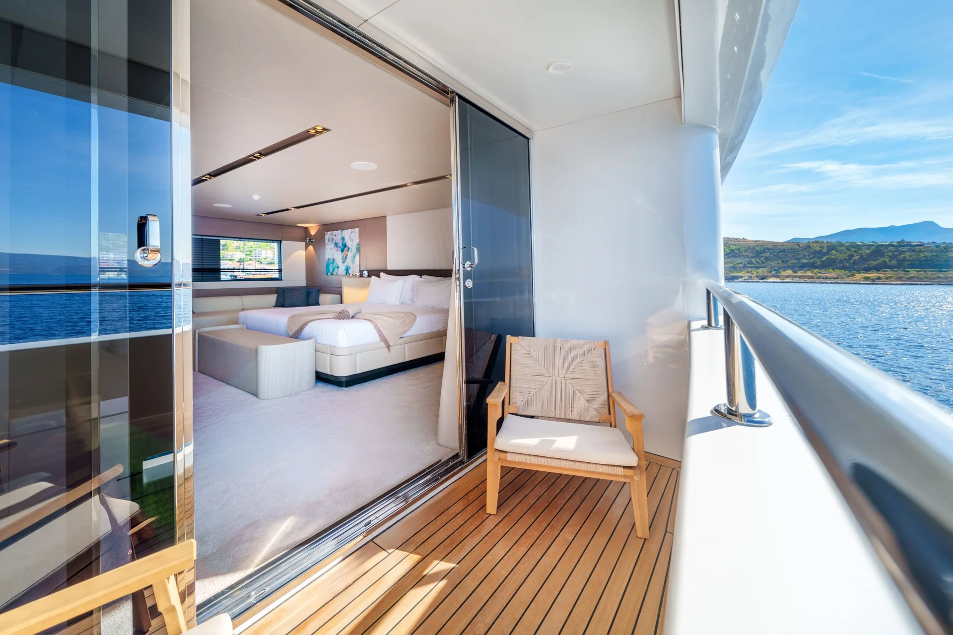 ORIY A modern yacht balcony with a wooden chair, glass doors open to a bright bedroom with a large bed, white décor, and a view of the sea and hills under a blue sky.