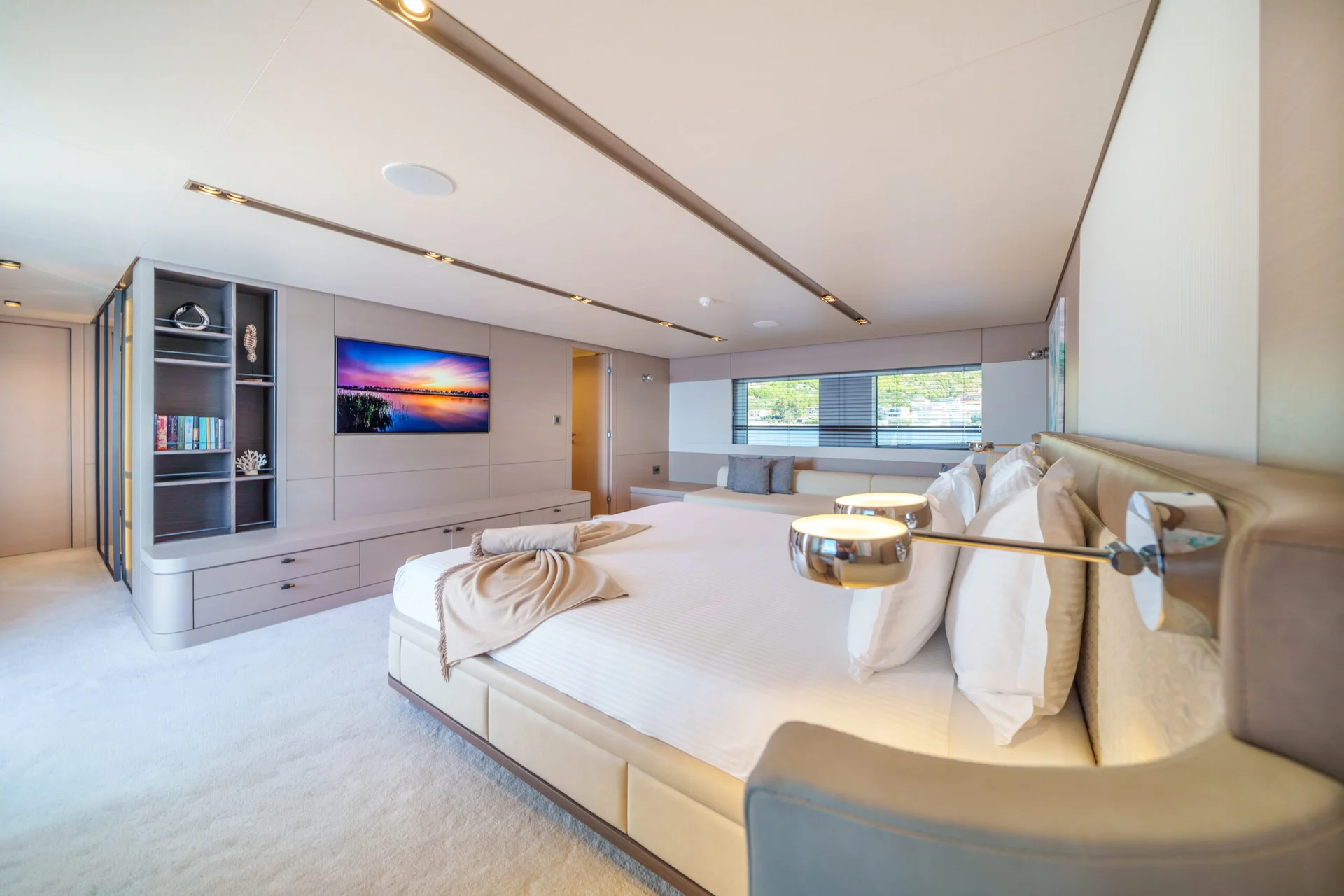 ORIY Spacious, modern bedroom with a large bed, neutral colors, built-in shelves, a wall-mounted TV displaying a sunset, and large windows letting in natural light.