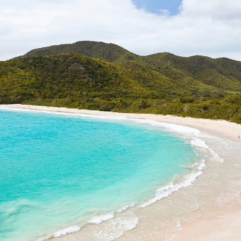 A turquoise ocean curves along a sandy beach with gentle waves; lush green hills rise in the background under a partly cloudy sky.