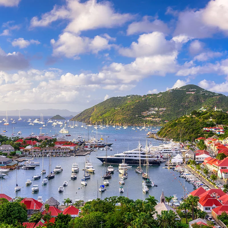 Colorful harbor town with red-roofed buildings, numerous boats and yachts on blue water, green hills in the background under a partly cloudy sky.