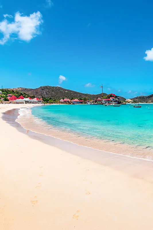 A tropical beach with white sand, clear turquoise water, gentle waves, and a few boats near the shore. Colorful tents and green hills are in the background under a bright blue sky with scattered clouds.