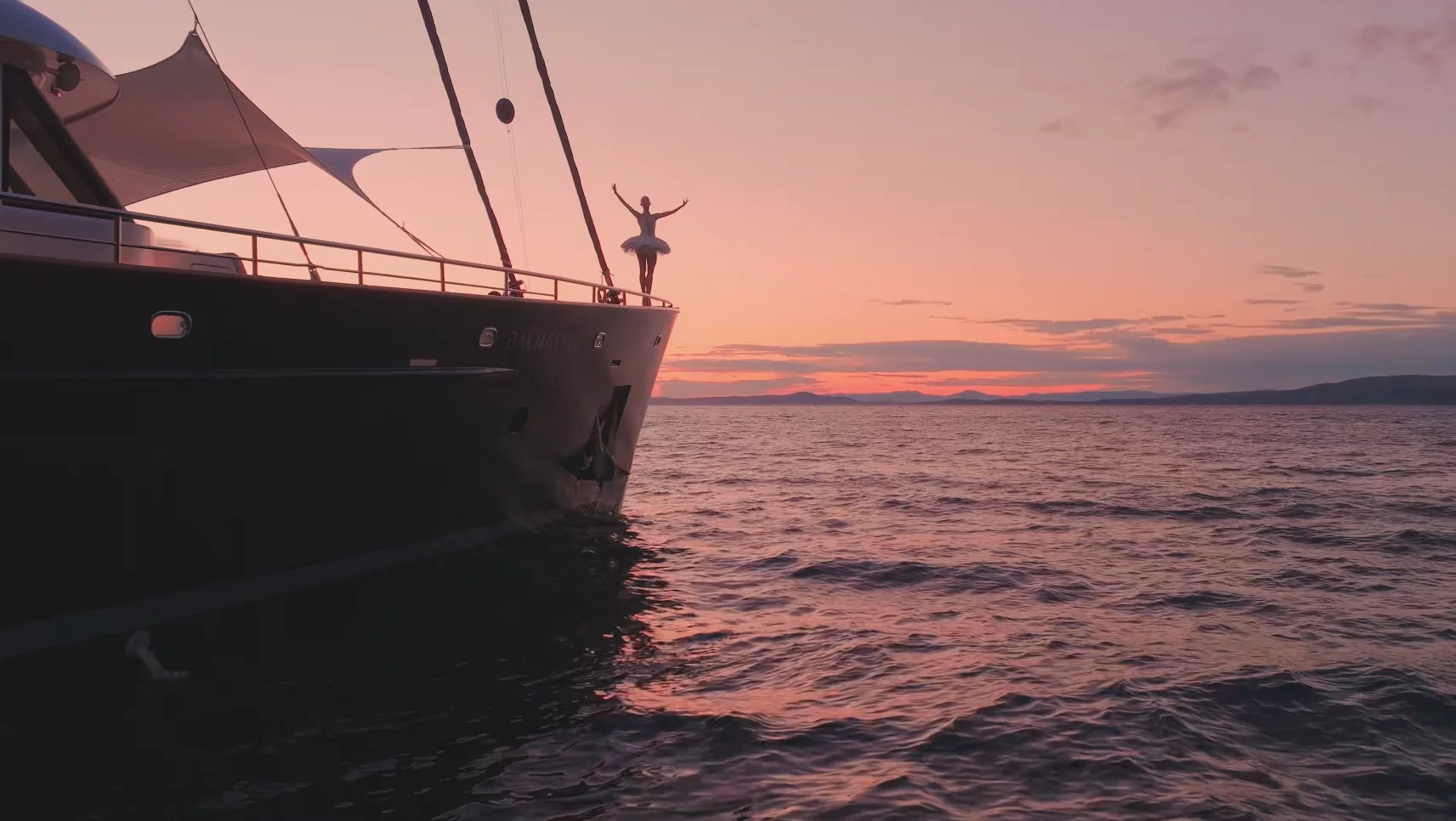 A person stands with arms raised on the bow of a yacht at sunset, overlooking calm ocean waters with orange and pink hues in the sky.