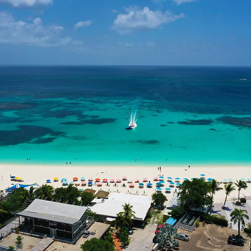 Aerial view of a tropical beach with turquoise water, colorful umbrellas, people on the sand, and a boat in the sea under a clear, sunny sky.