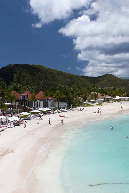 A tropical beach with white sand, turquoise water, people relaxing, and red-roofed buildings surrounded by green hills under a partly cloudy sky.