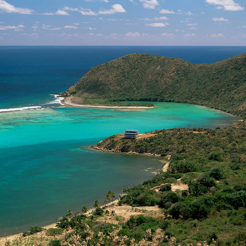 A vivid turquoise bay meets a lush, hilly coastline under a bright sky with scattered clouds. A small building sits near the shore, surrounded by greenery and sandy paths.