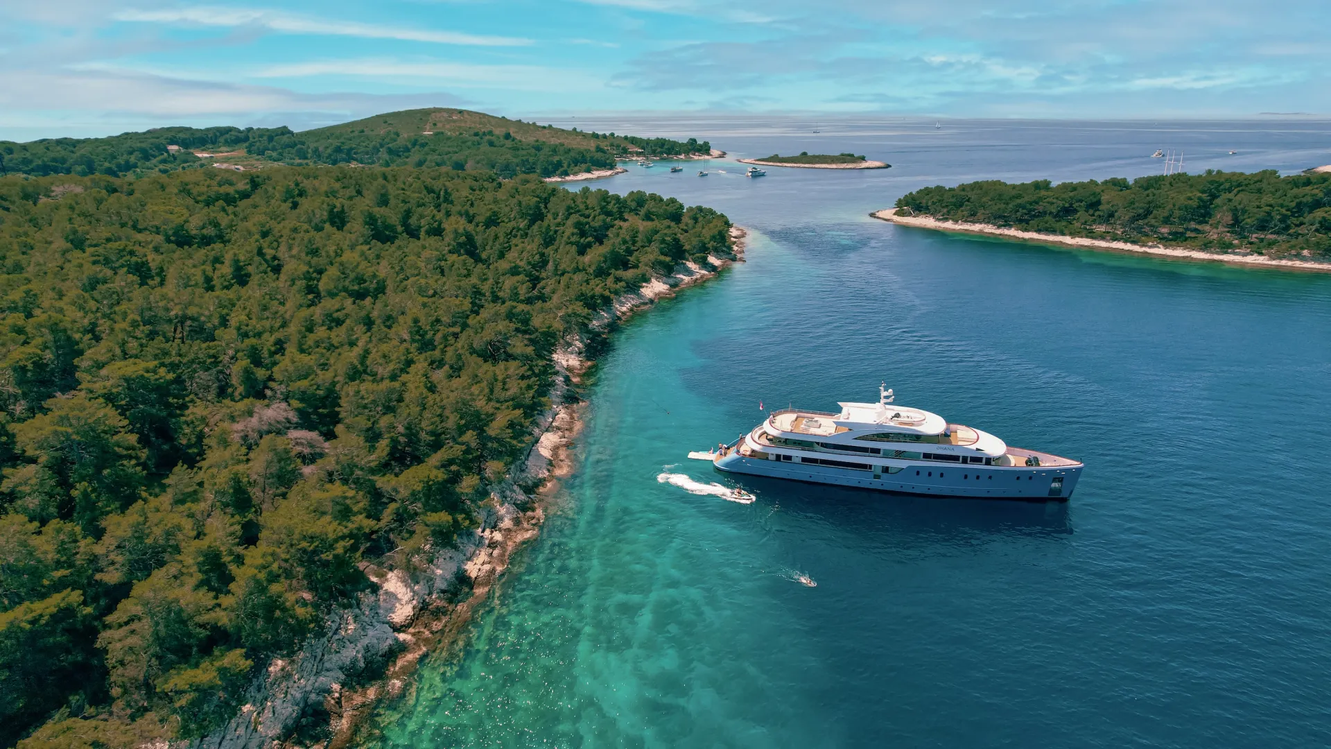 Un gran yate blanco navega por aguas azules y cristalinas cerca de una isla de exuberante vegetación y costas rocosas, bajo un cielo luminoso y parcialmente nublado.