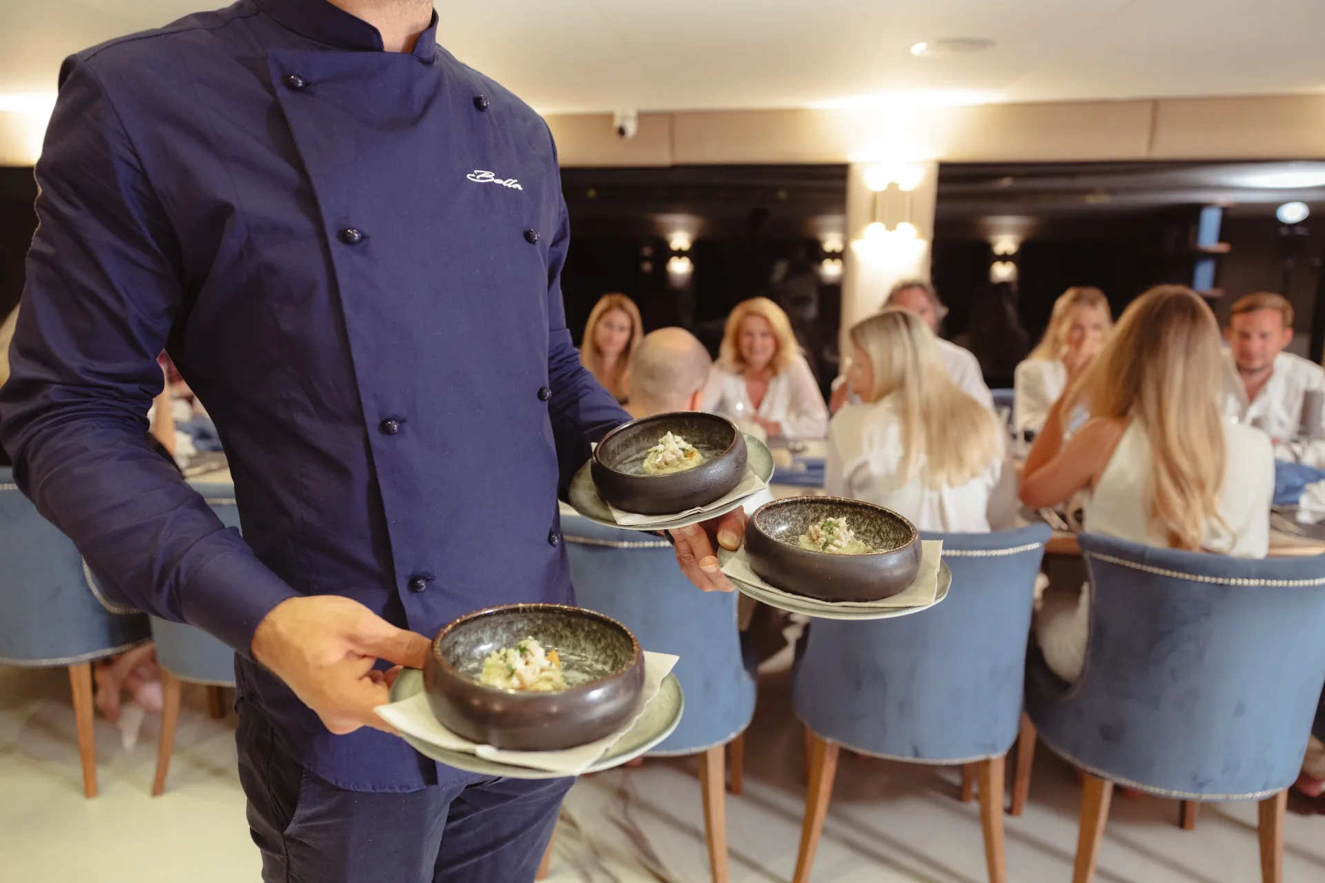 Un camarero, vestido con una chaqueta azul de chef, lleva tres cuencos de comida a un grupo de personas sentadas a una mesa de un restaurante.
