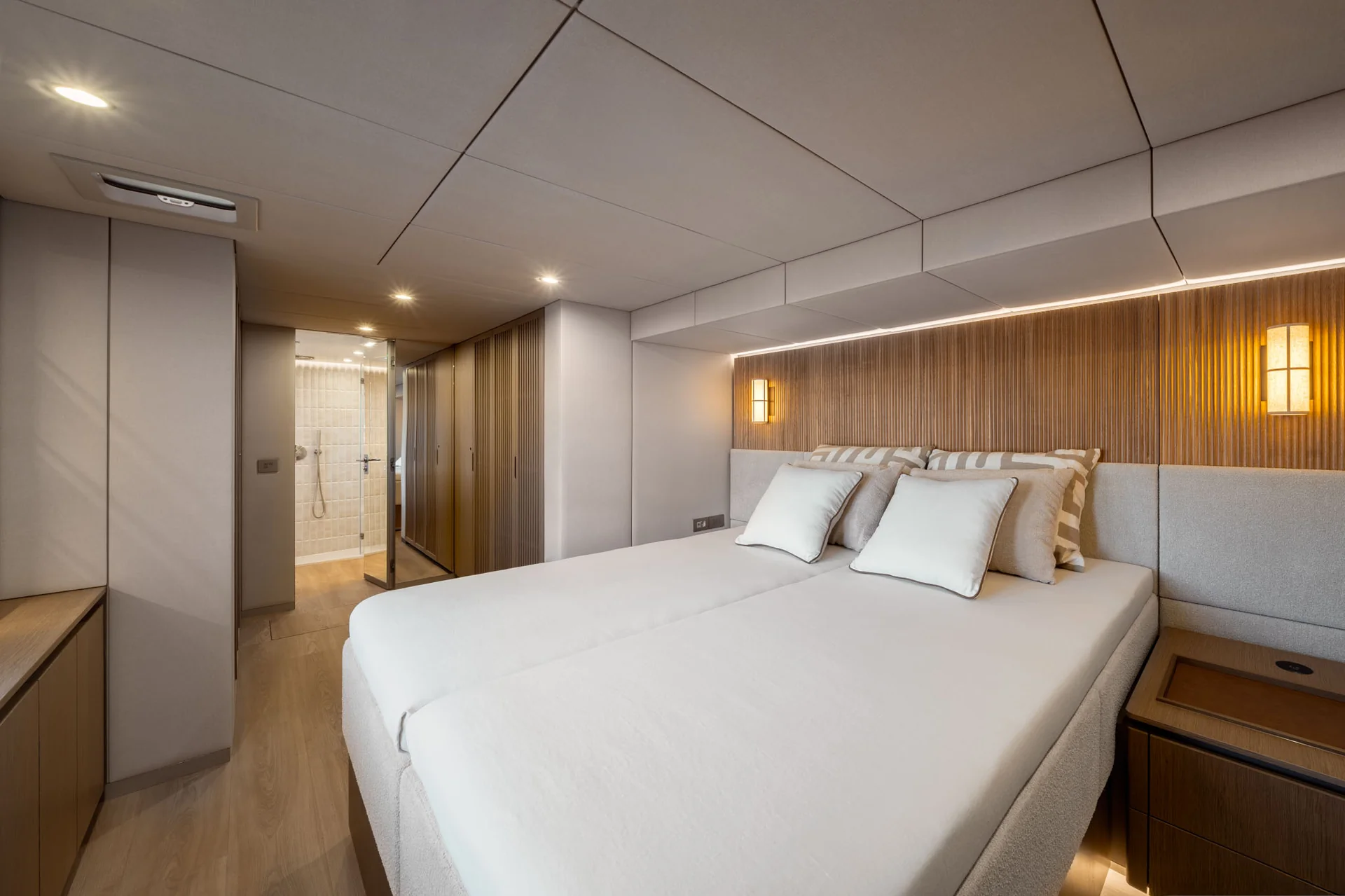 Modern yacht cabin featuring a double bed with white linens against a slatted wooden headboard and an adjacent private bathroom.