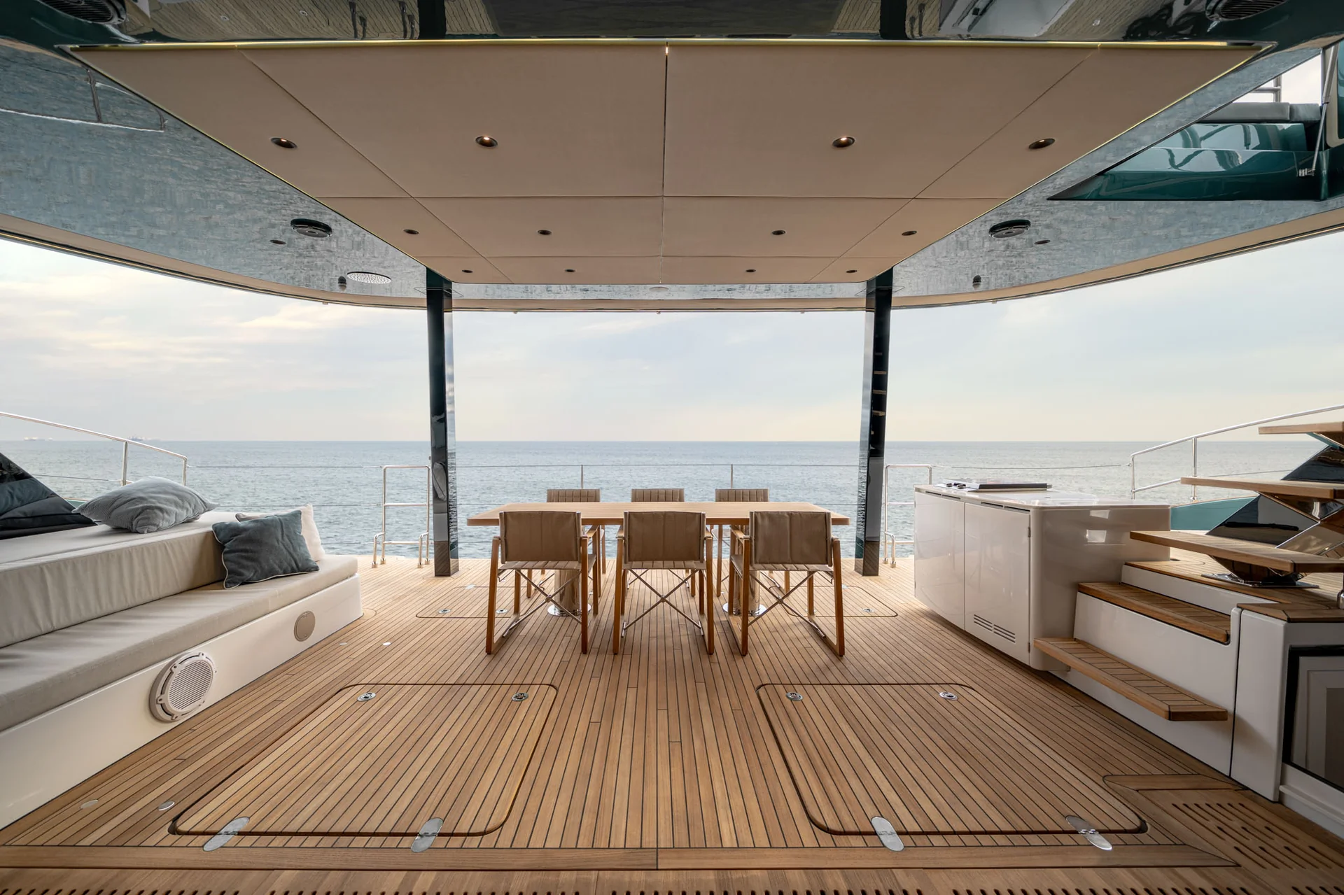 The open-air aft deck of a yacht features a dining table with six chairs, a cushioned lounge area, and a wooden floor, all overlooking the ocean.