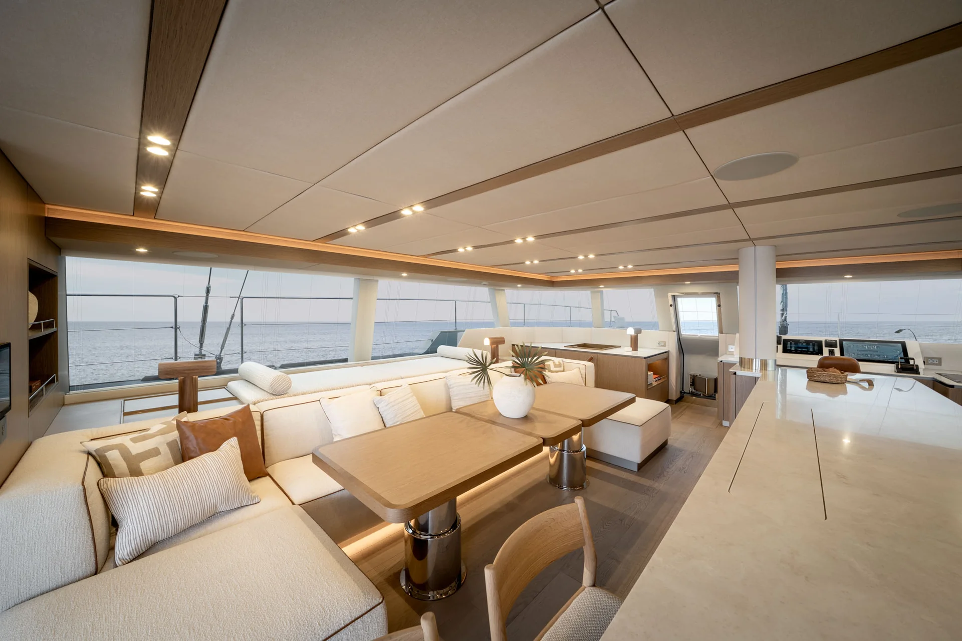 A modern yacht interior featuring a white L-shaped sofa, two wooden dining tables, and a large countertop, with expansive windows overlooking the ocean.