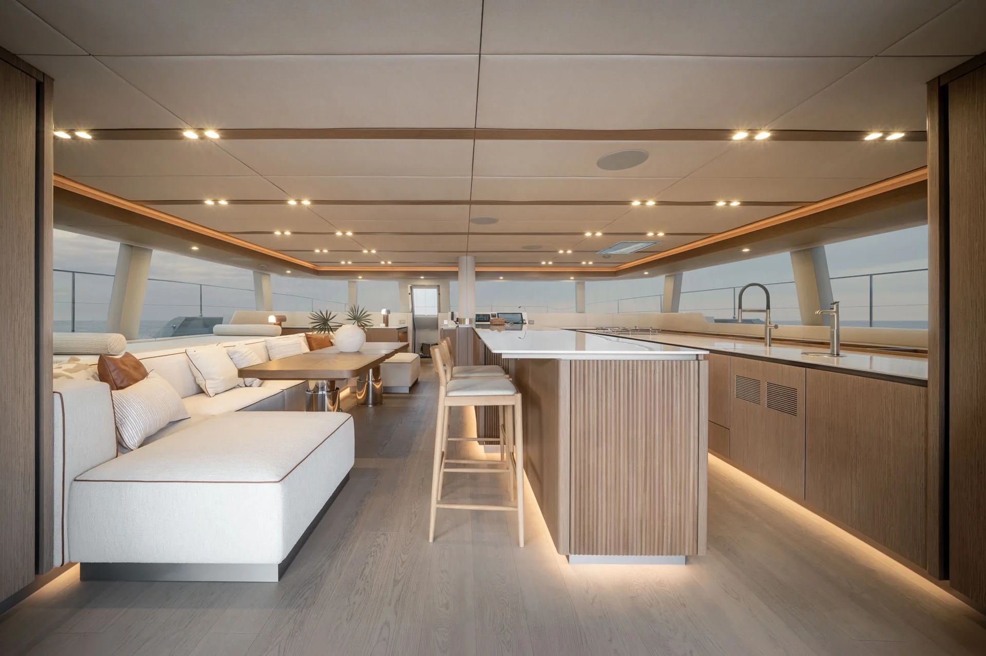 Interior of a yacht featuring a lounge area with an L-shaped sofa and dining table, and a kitchen island with bar stools.