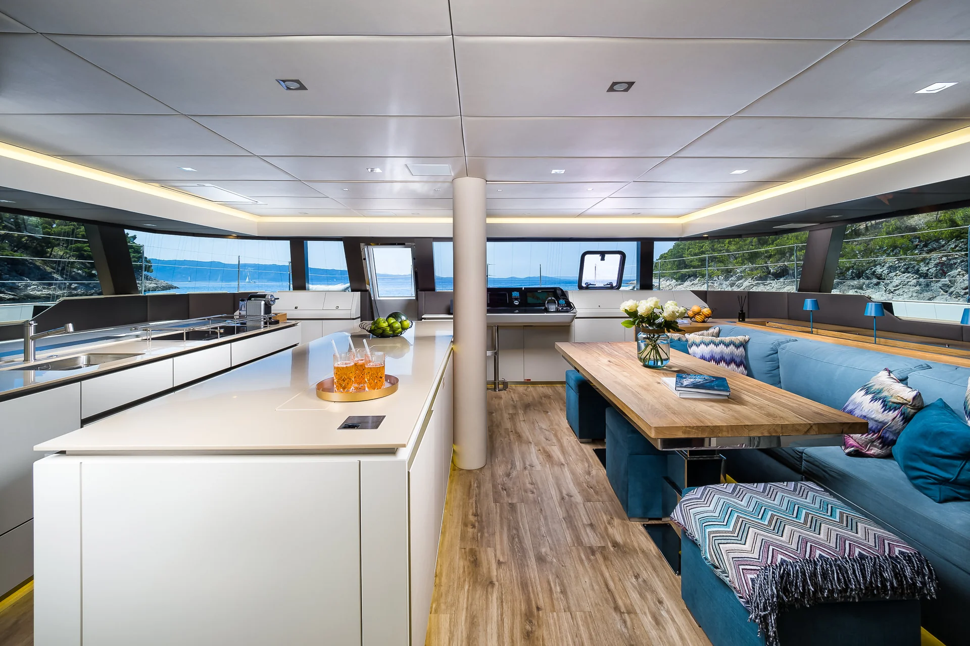 Modern yacht interior featuring a white kitchen island, a wooden dining table with blue upholstered seating, and large windows with views of the sea.