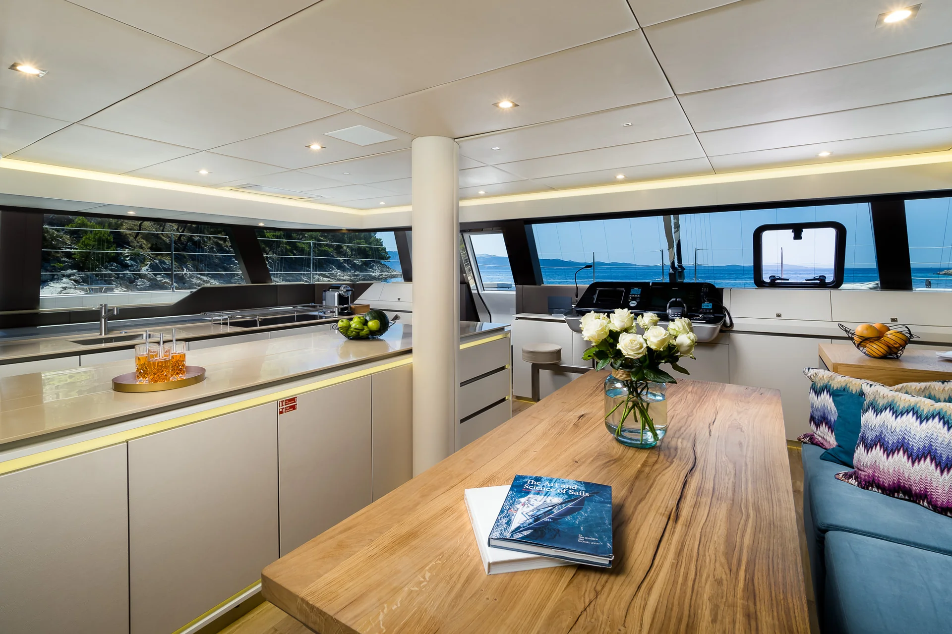 The interior of a modern yacht featuring a kitchen area, a large wooden dining table with a bouquet of white roses, and a blue cushioned bench, with expansive windows overlooking the sea.