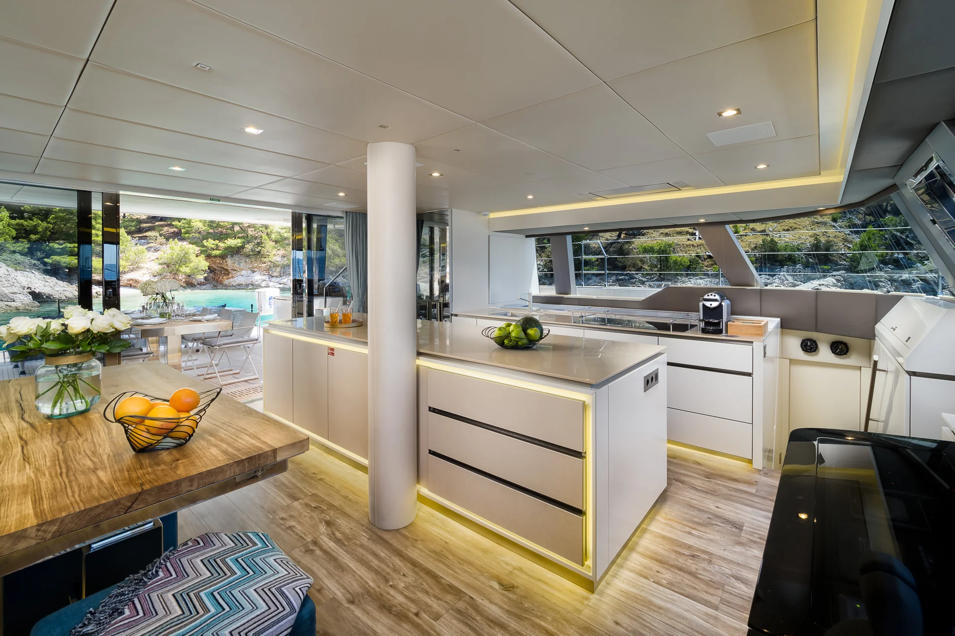 Modern kitchen and dining area on a yacht, featuring a large central island, wooden table, and sliding glass doors opening to a view of the water.