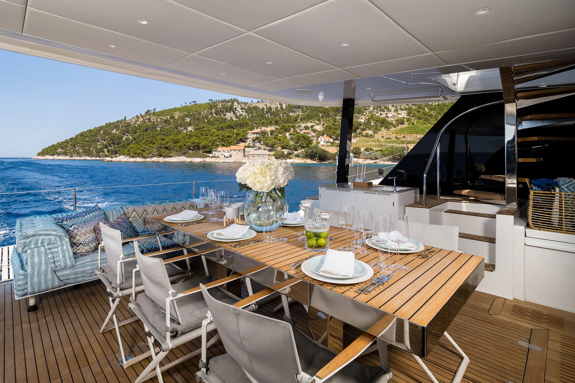 Outdoor dining area on a yacht deck with a wooden table set for six, featuring white plates, glassware, and a large floral centerpiece, overlooking a coastal landscape.