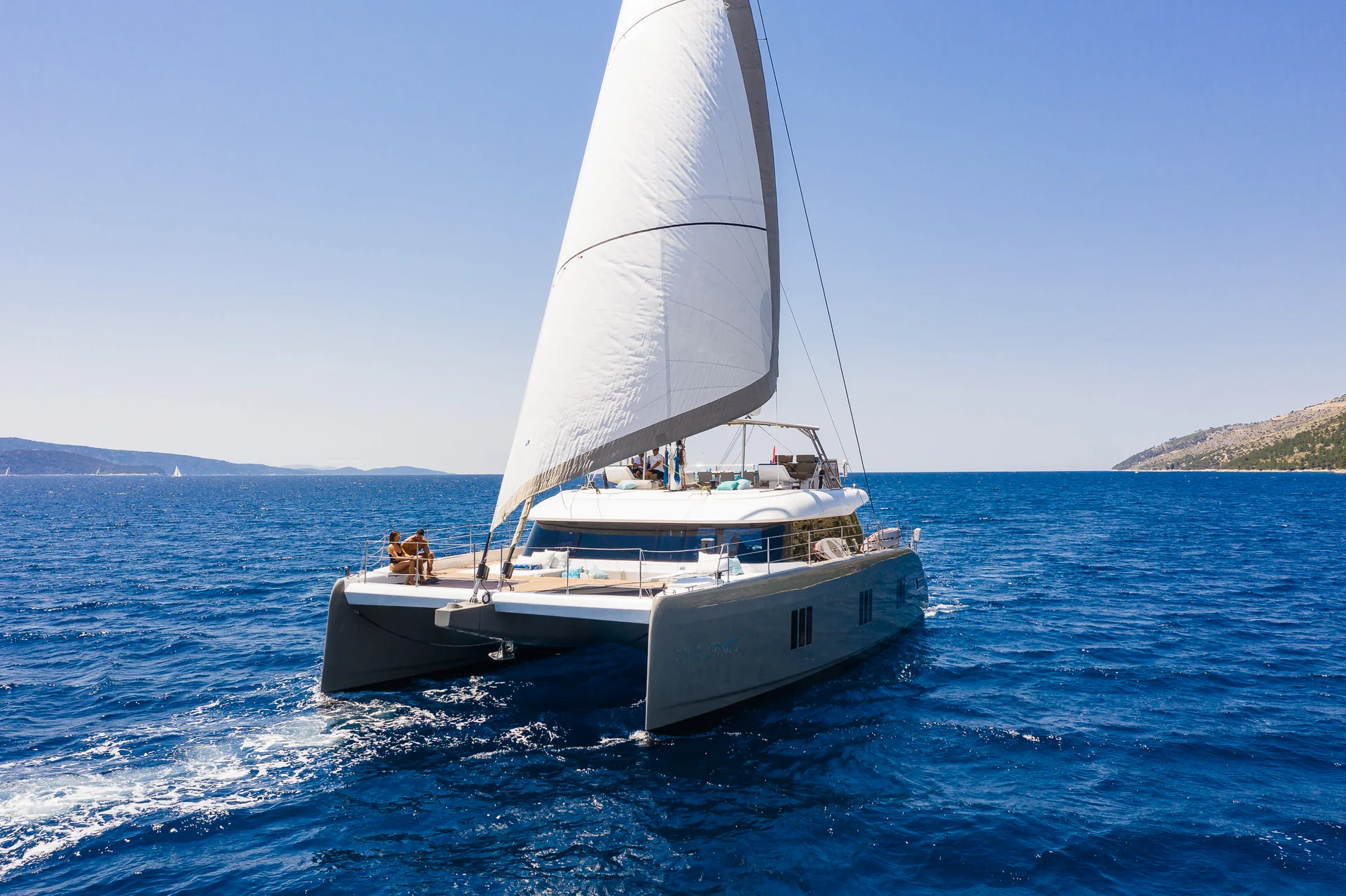 A catamaran with a white sail moving across the blue sea, with two people sitting on the front deck.