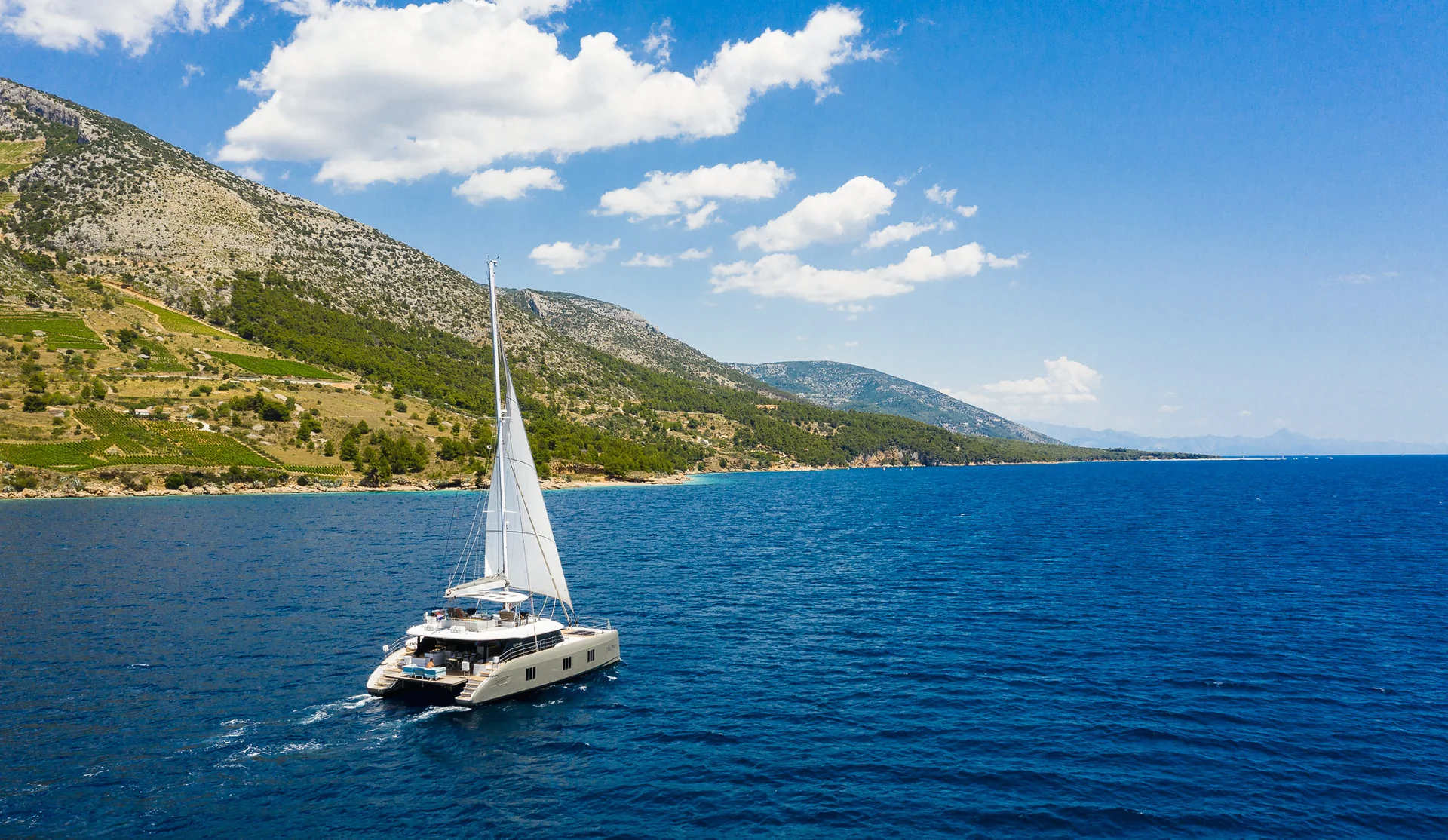 A catamaran sailboat cruising on blue water near a mountainous coastline.
