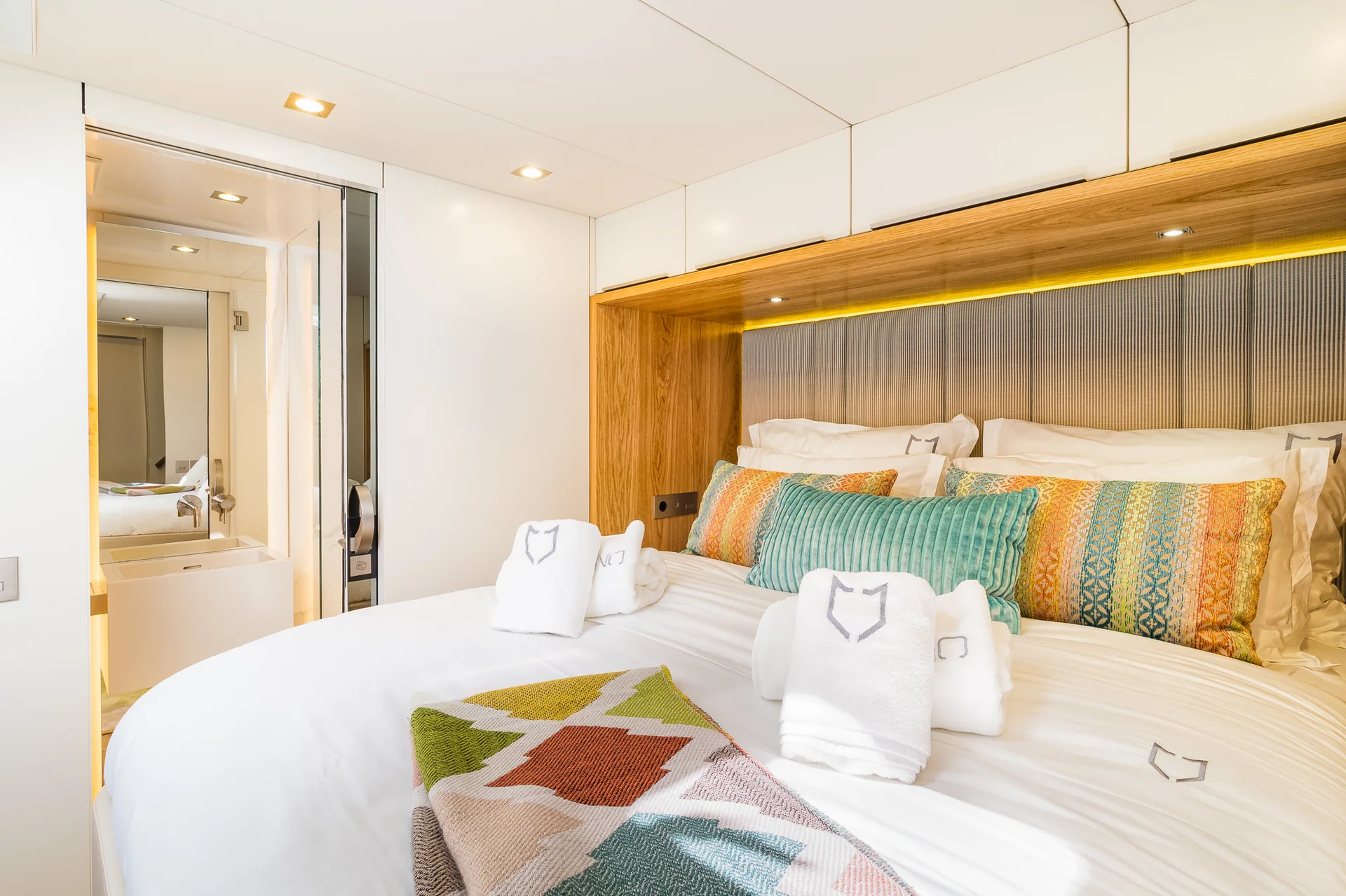 A neatly made bed with white linens, patterned pillows, and folded towels in a modern cabin, with an adjacent view of a bathroom.