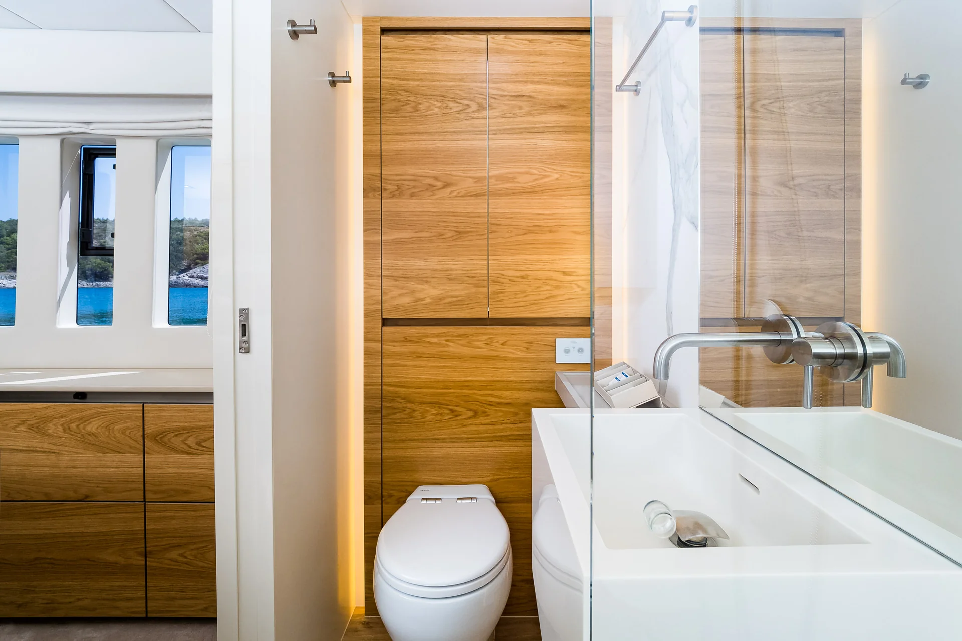 A modern bathroom with a white toilet, a floating sink, and wooden cabinetry, featuring a window view of the ocean.