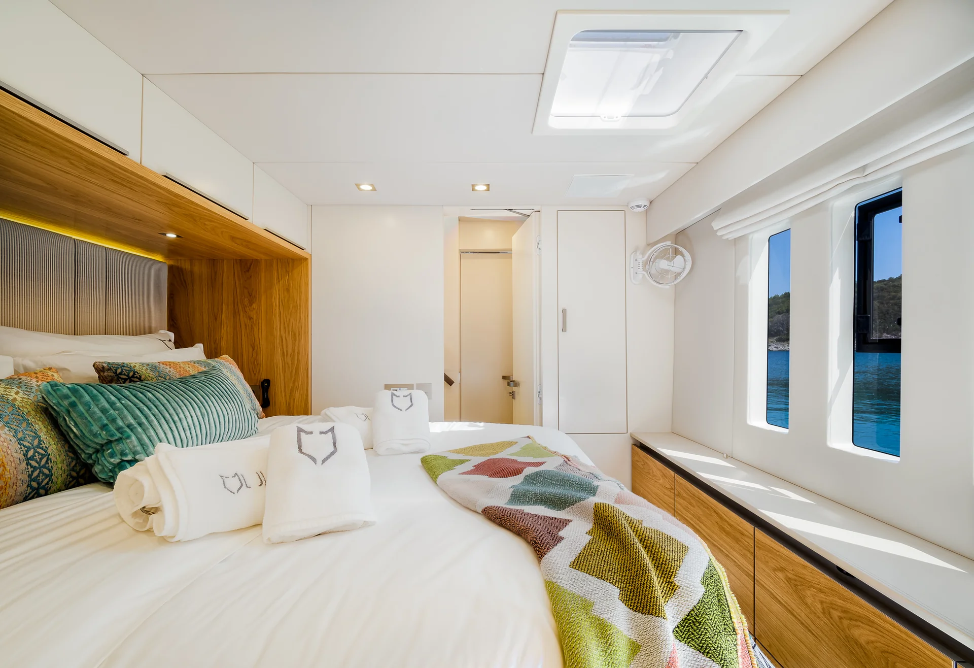 A yacht bedroom featuring a queen bed with white linens, logo-embroidered towels, and a colorful patterned throw blanket, with two windows looking out onto the sea.